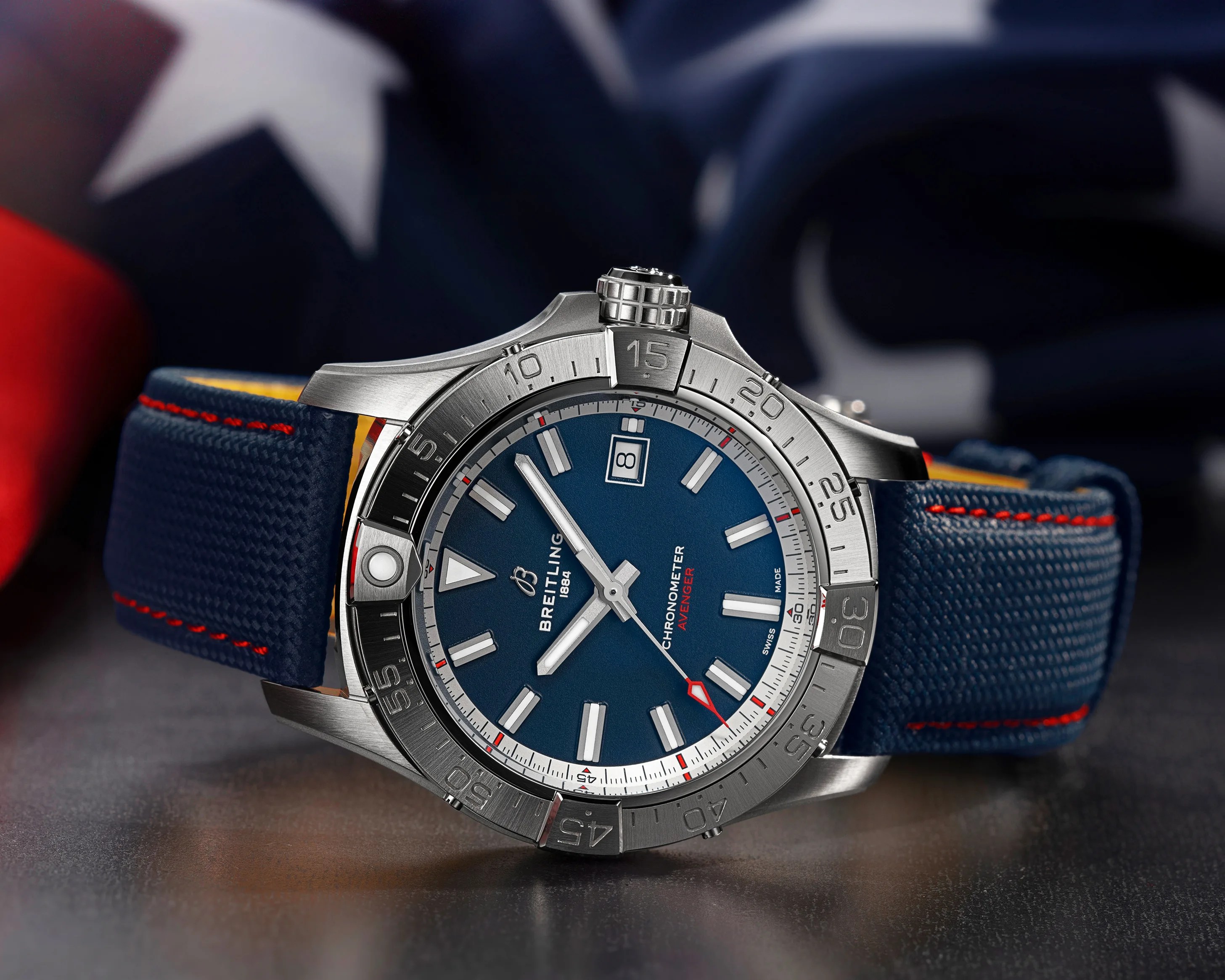 Silver Breitling Avenger chronometer watch with blue dial and blue fabric strap with red stitching on a dark surface.