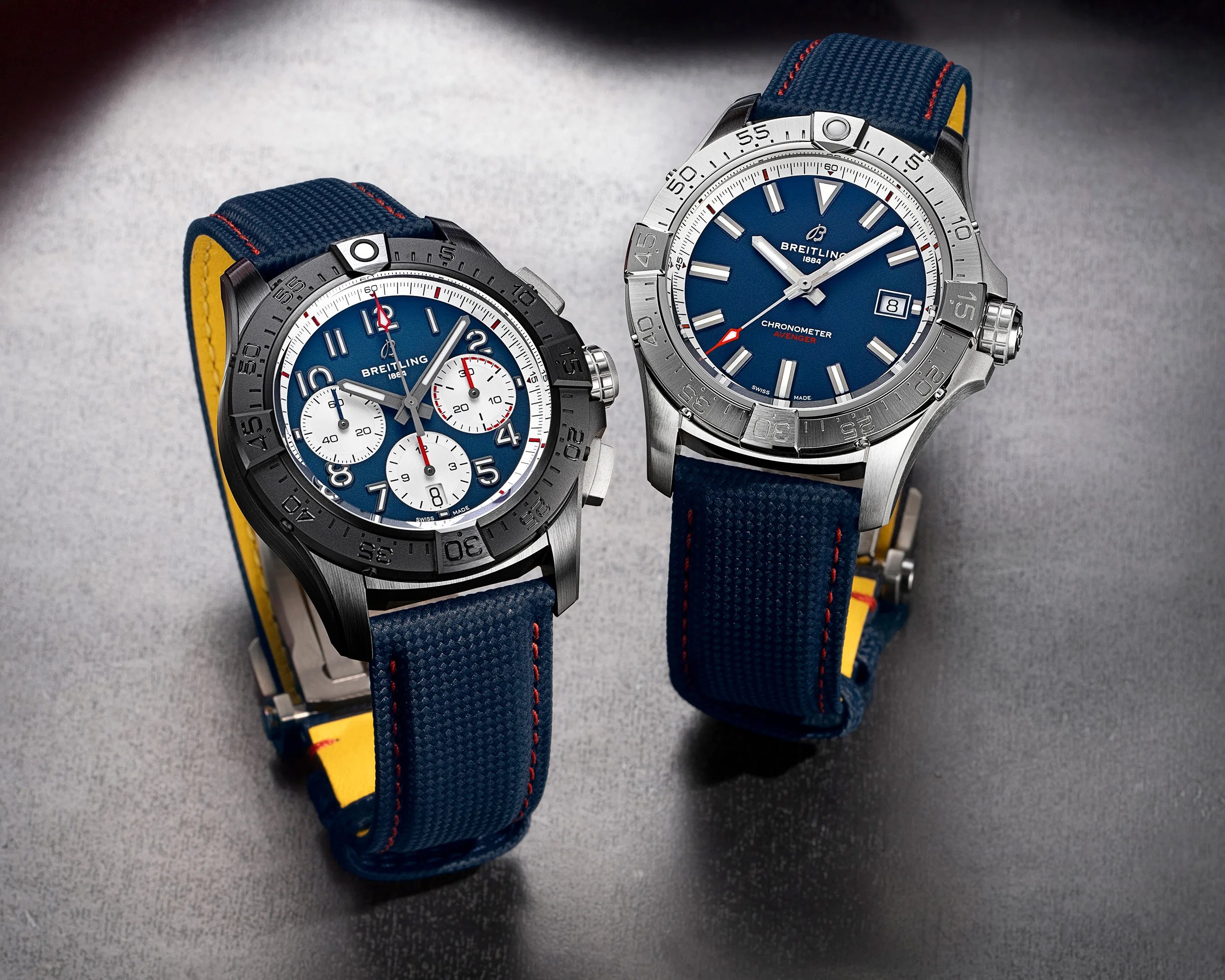 Two Breitling wristwatches with blue dials and blue fabric straps with yellow undersides on a gray surface.