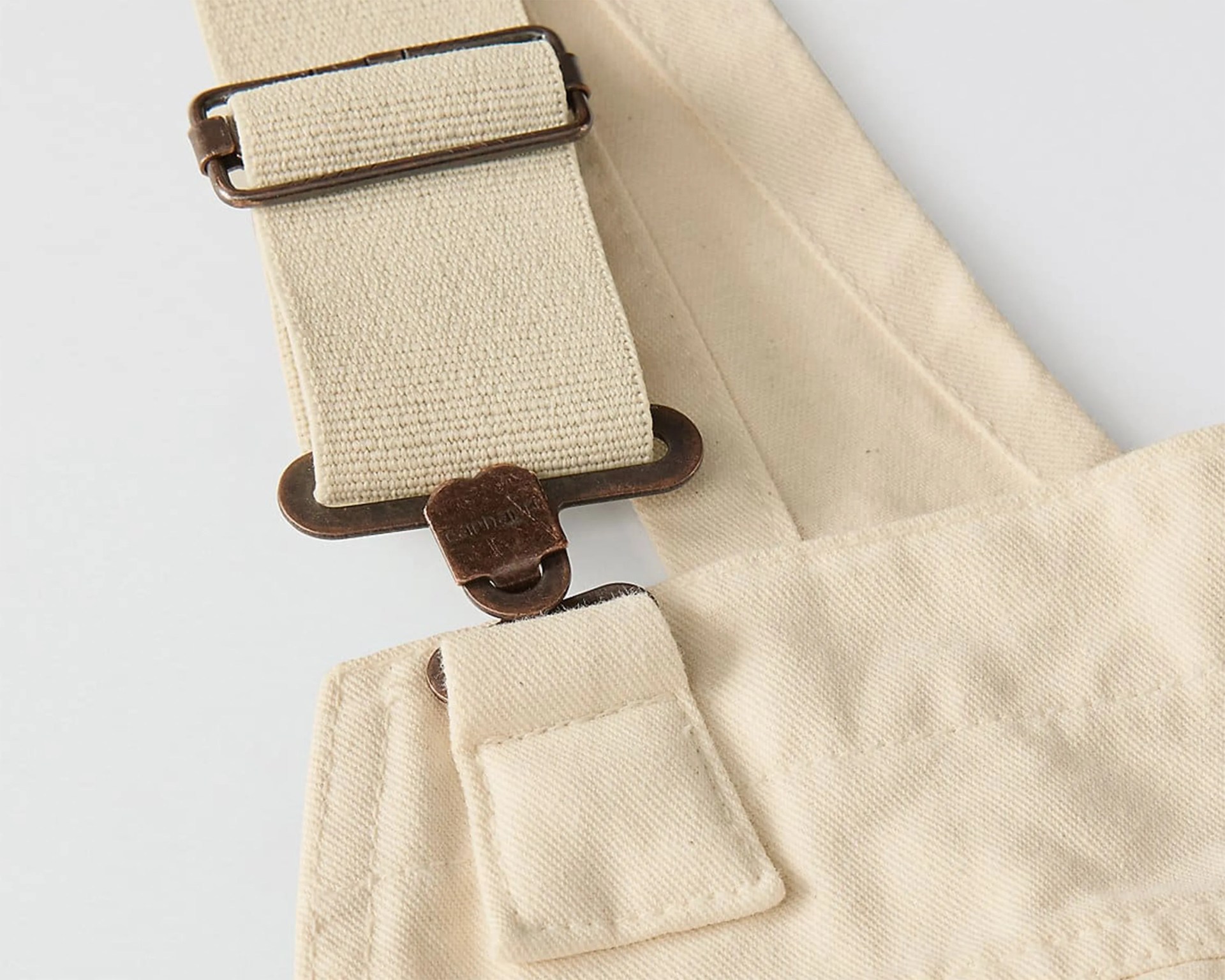 Close-up of beige canvas strap with antique brass buckle and stitching details on fabric.