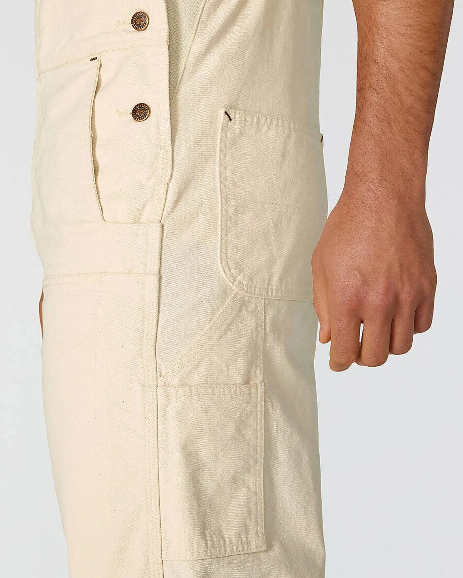 Close-up of beige pants with metal buttons and multiple pockets, worn by a person with a visible hand.