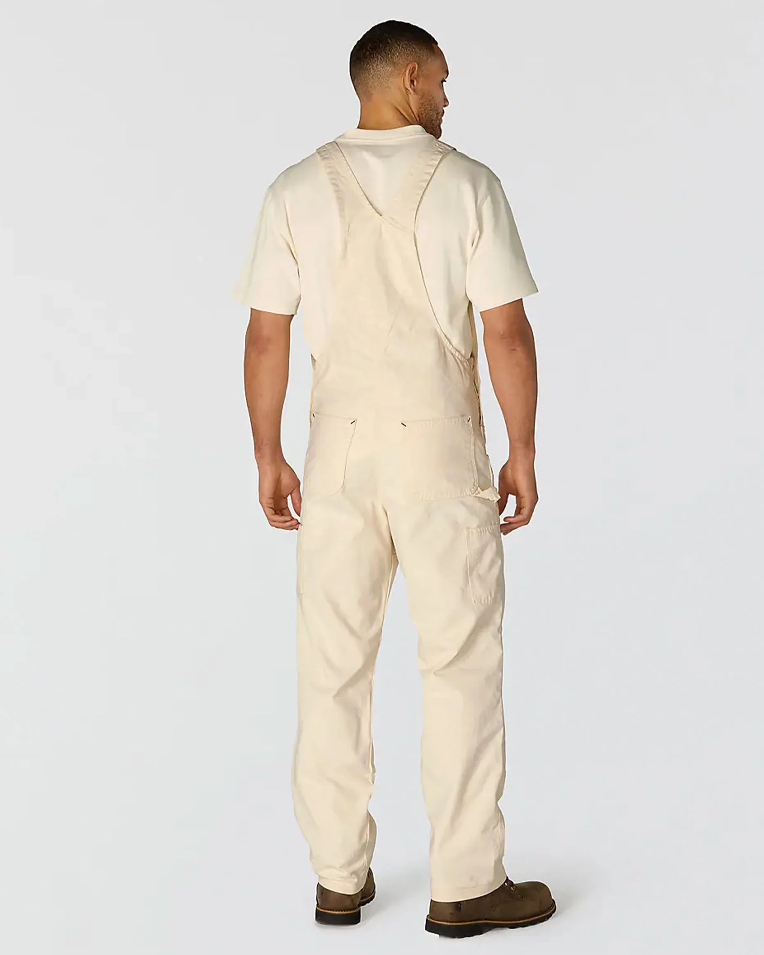Man wearing beige canvas overalls and a matching short-sleeve shirt with brown work boots, shown from the back.