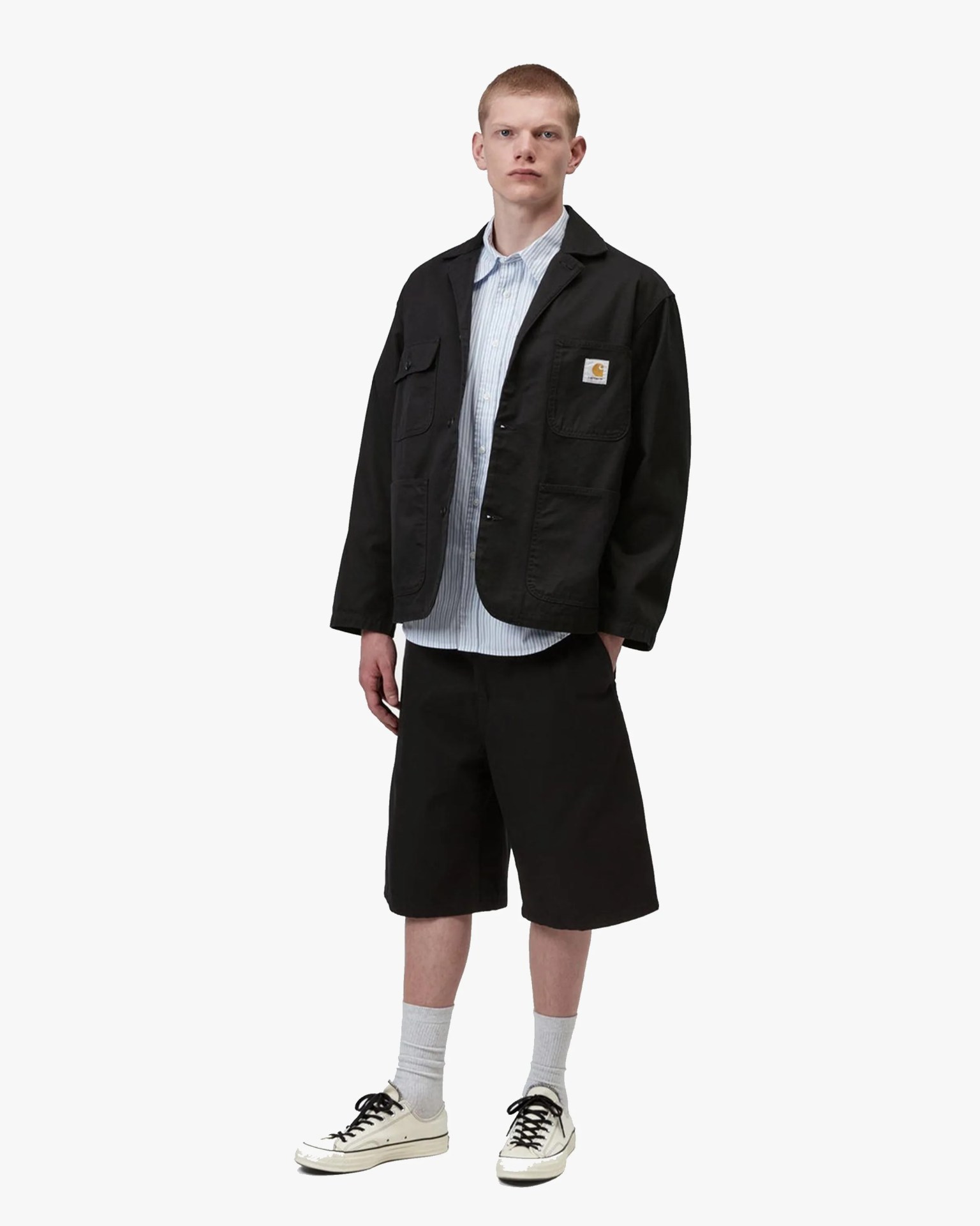 Black Carhartt chore jacket with patch logo on chest pocket worn over a light blue striped button-up shirt.