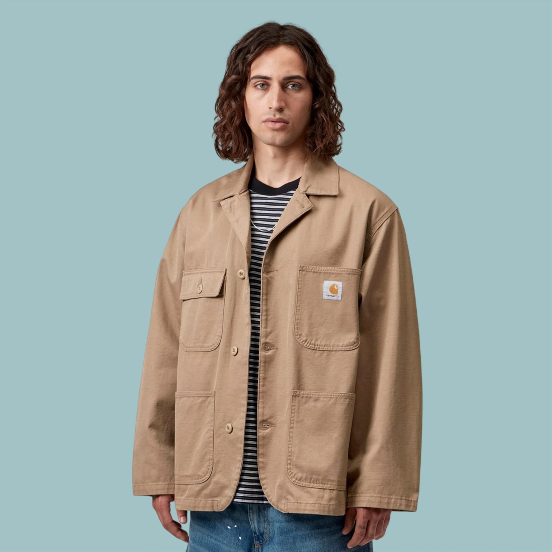 Beige Carhartt jacket with buttoned flap pockets worn over a black and white striped shirt.