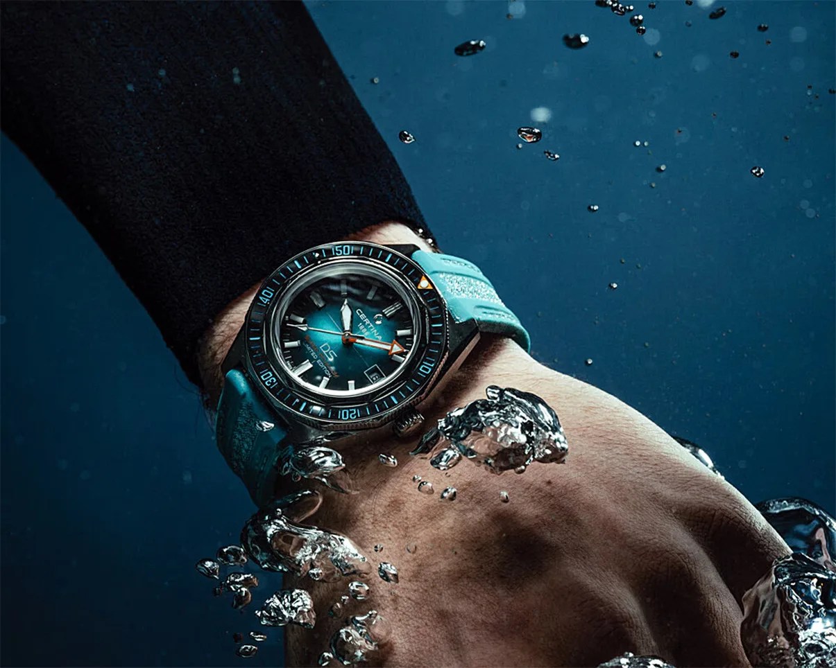 Wristwatch with blue dial and matching blue strap worn underwater with bubbles around.