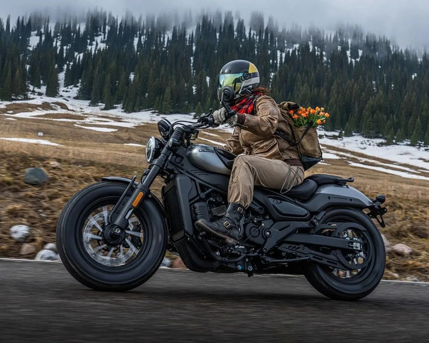Person wearing a helmet and brown jacket riding a black motorcycle on a road with snowy pine trees in the background.