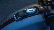 Close-up of a motorcycle fuel tank with a chrome fuel cap and a black strap with a red stripe.