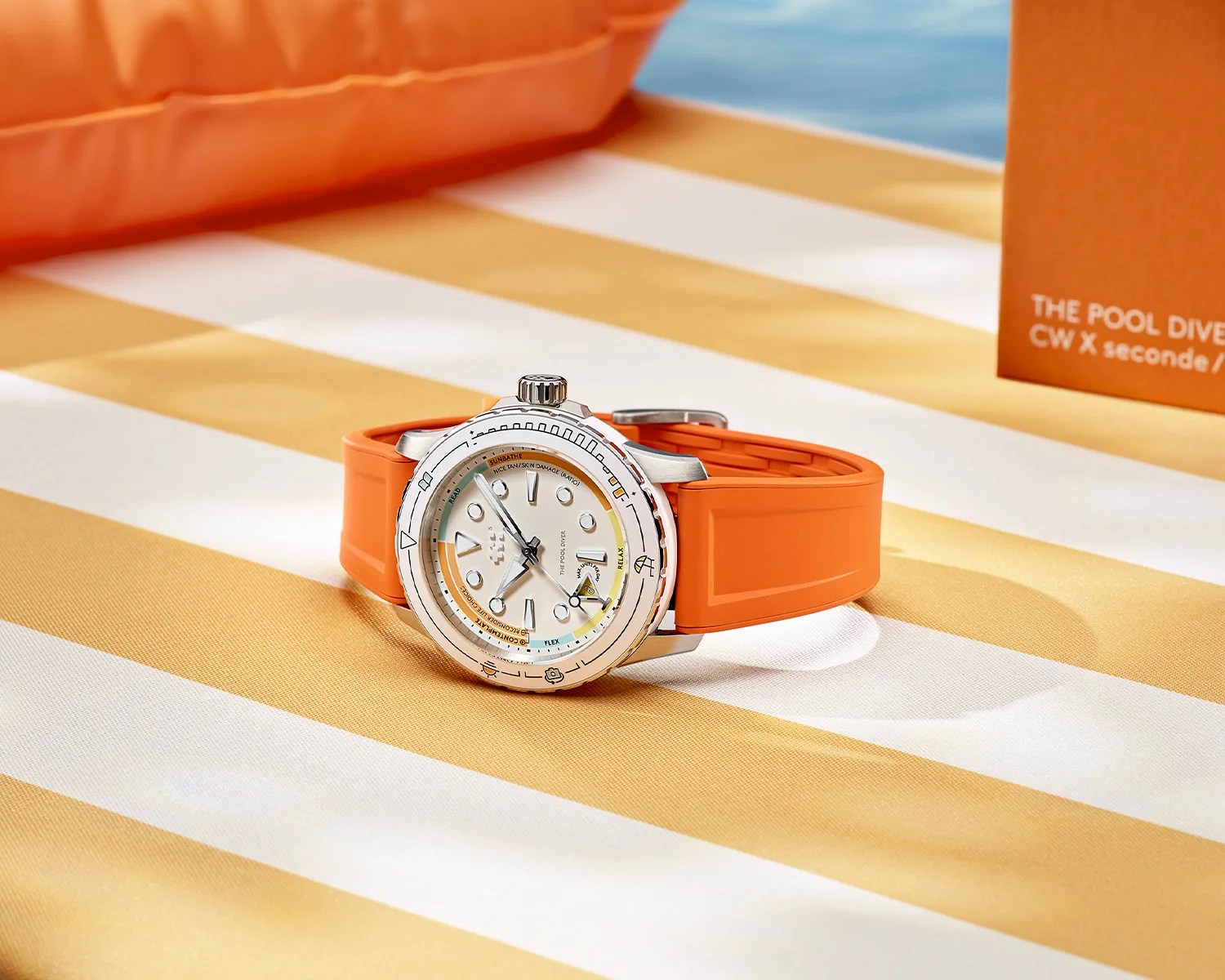 Silver dive watch with white dial and orange rubber strap on yellow and white striped fabric.