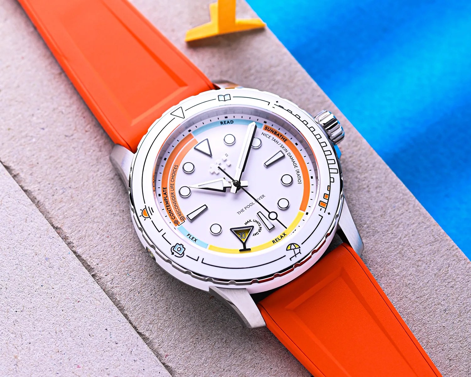 Silver watch with white dial, orange and yellow accents, and bright orange rubber strap on a stone surface near blue water.
