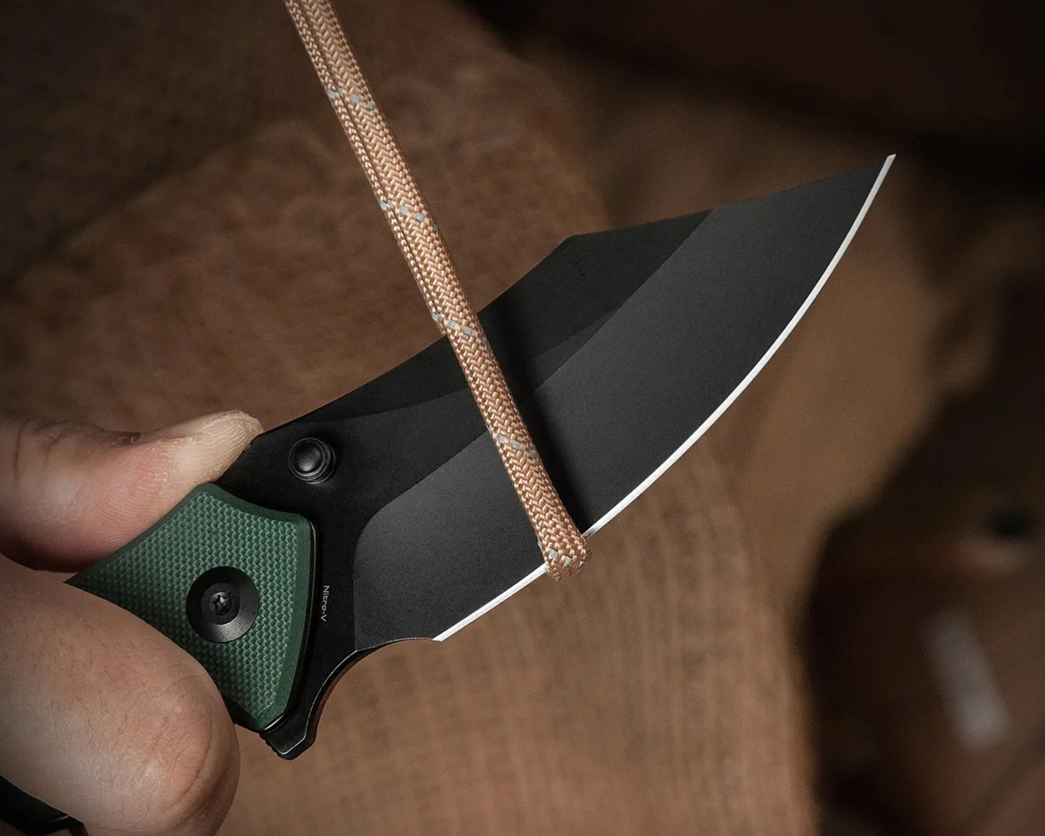 Black folding knife with a green textured handle cutting a tan rope.