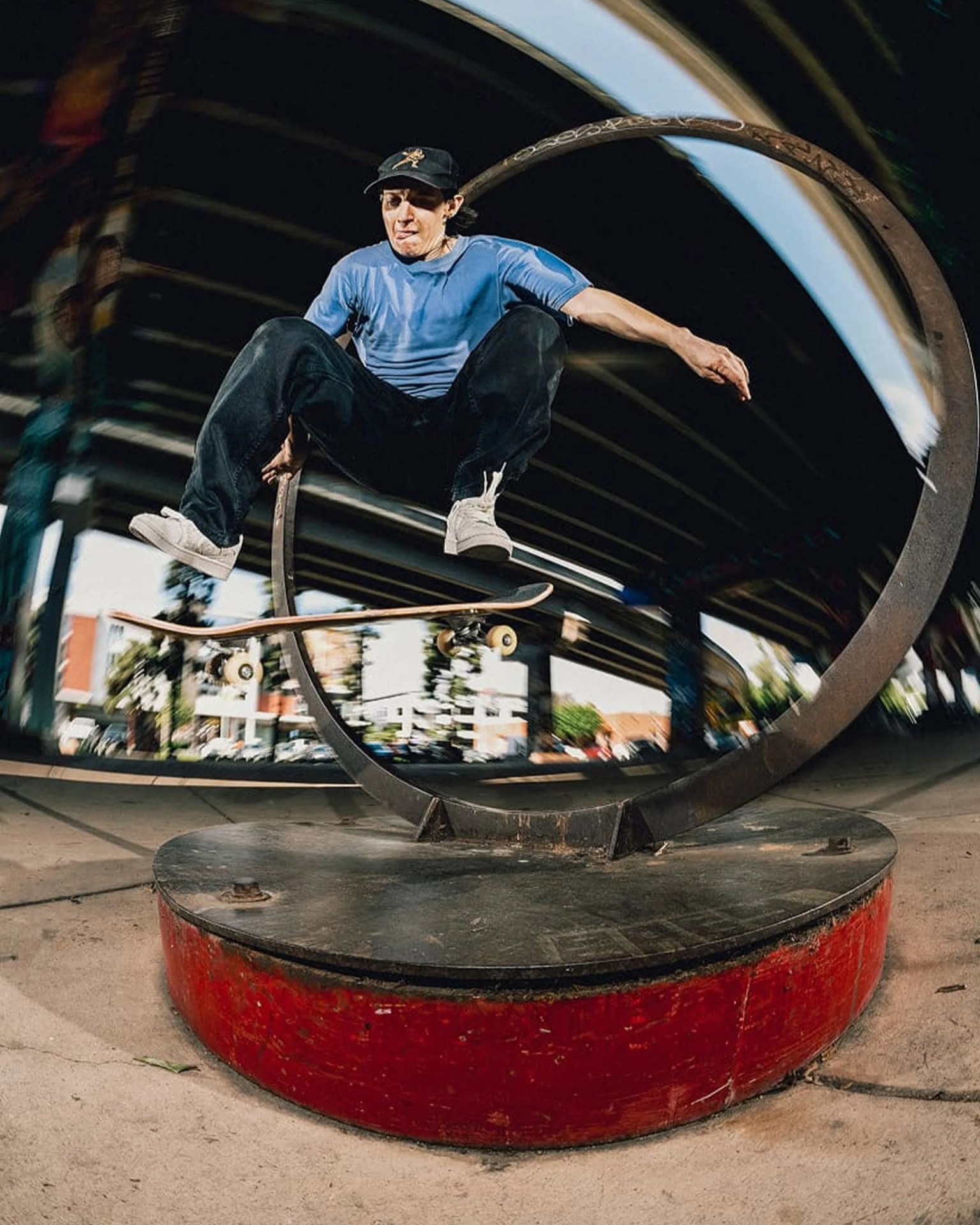 Skateboarder in a blue shirt and black pants performing a trick over a circular metal sculpture on a red base.