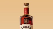 Close-up of the top half of a Coors whiskey bottle with a wooden cap and red label on a beige background.