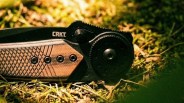 Black and brown CRKT folding knife with textured handle resting on mossy ground.