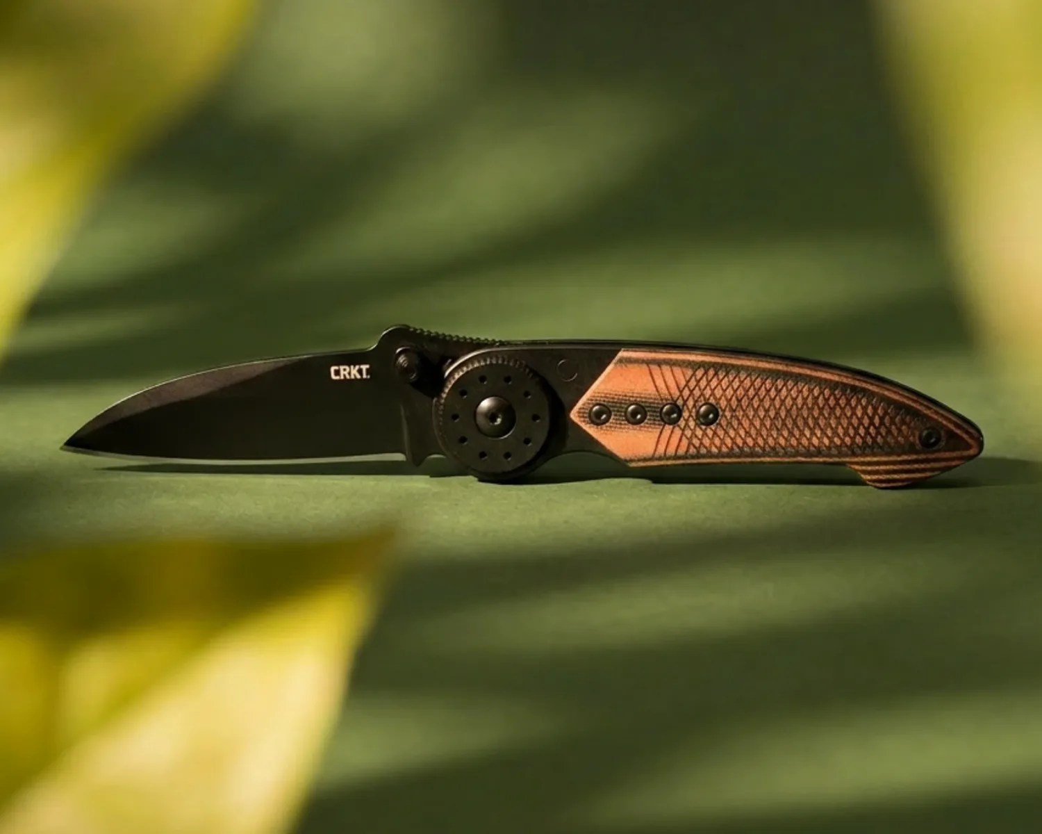 Black folding knife with wooden textured handle and CRKT logo on the blade, set on a green surface.