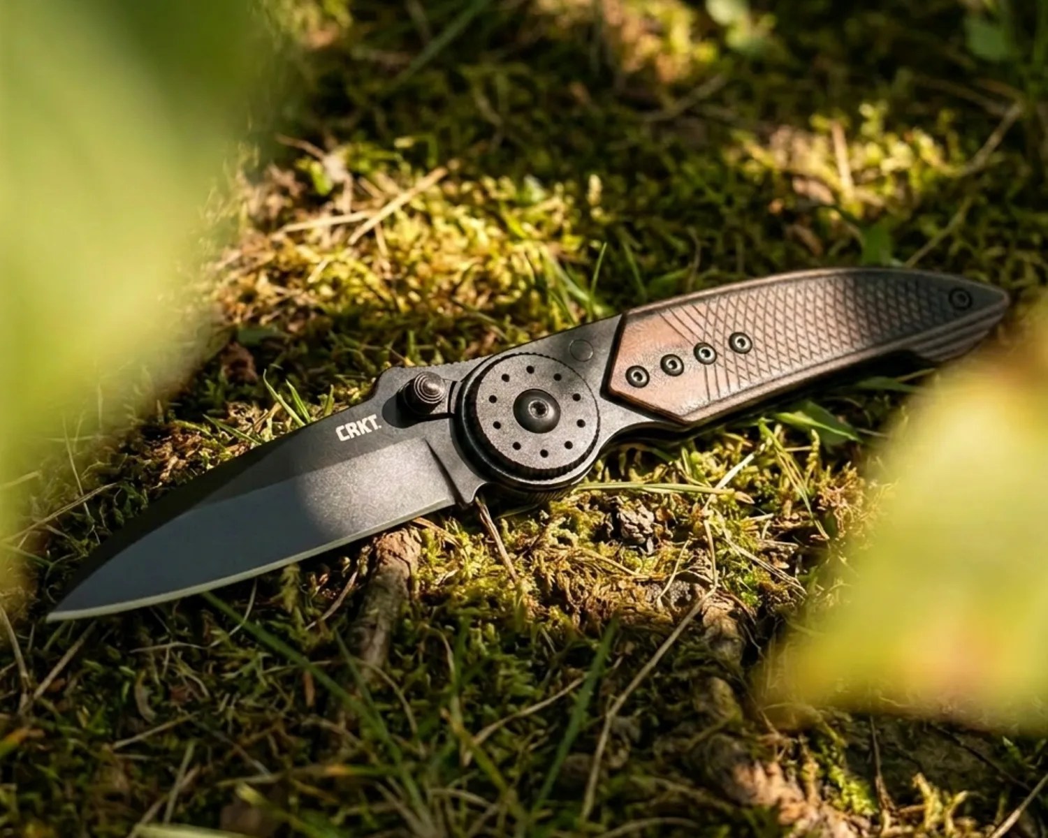 Black folding knife with textured brown handle and CRKT logo on blade lying on mossy ground.
