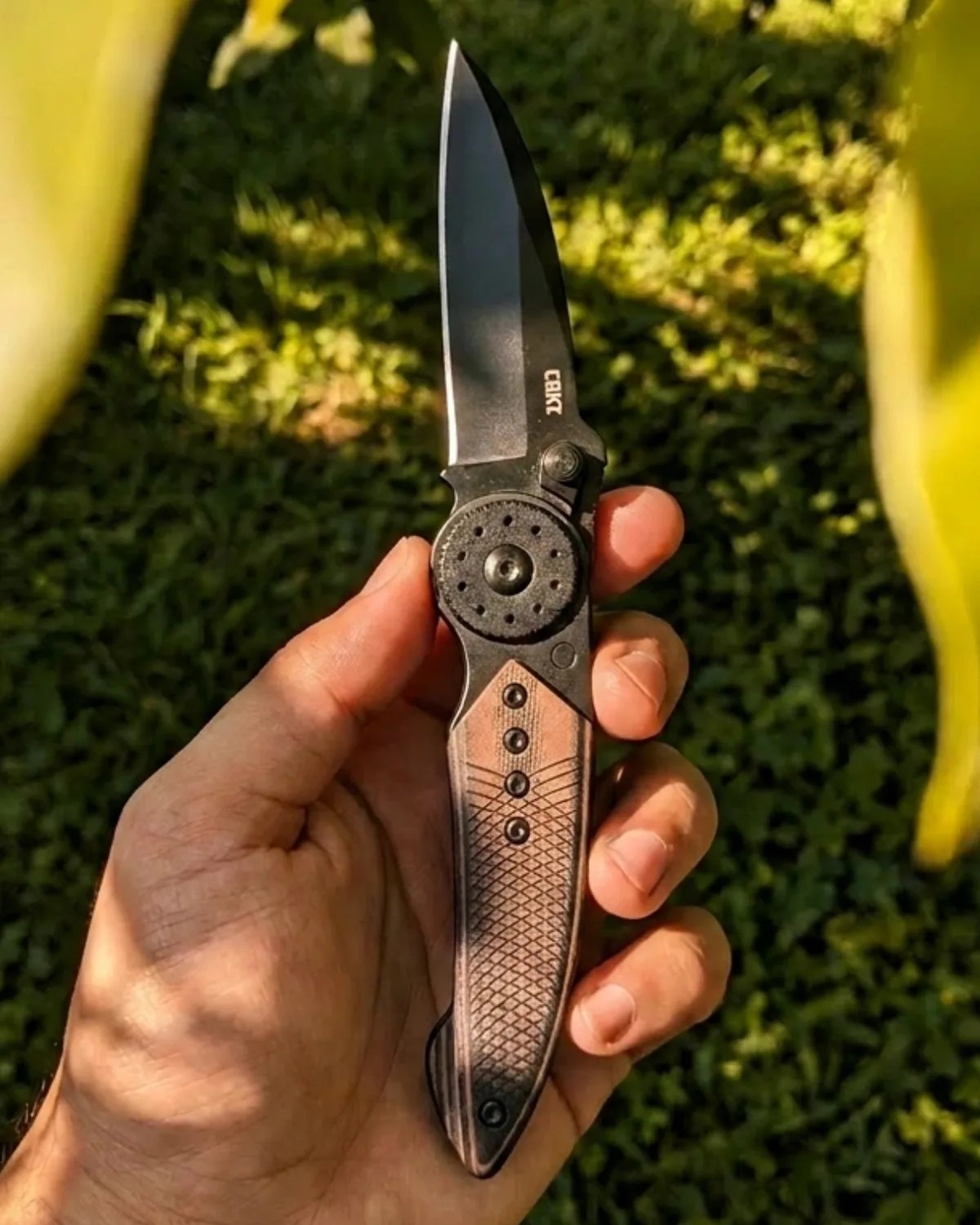 Hand holding a folding knife with a black blade and textured brown handle outdoors.