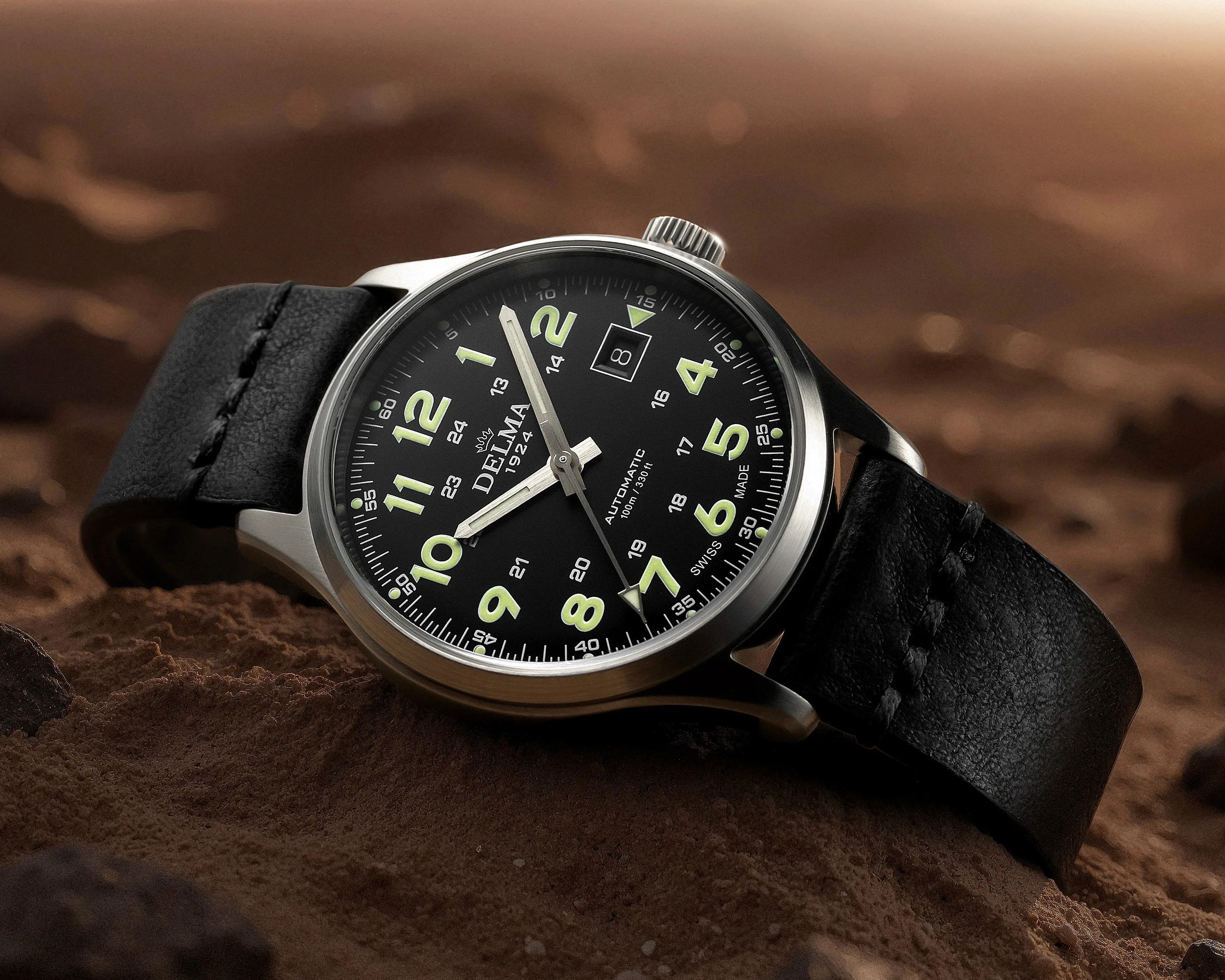 Close-up of a Delma 1924 automatic wristwatch with black dial, luminous green numerals, date window, and black leather strap on brown textured surface.