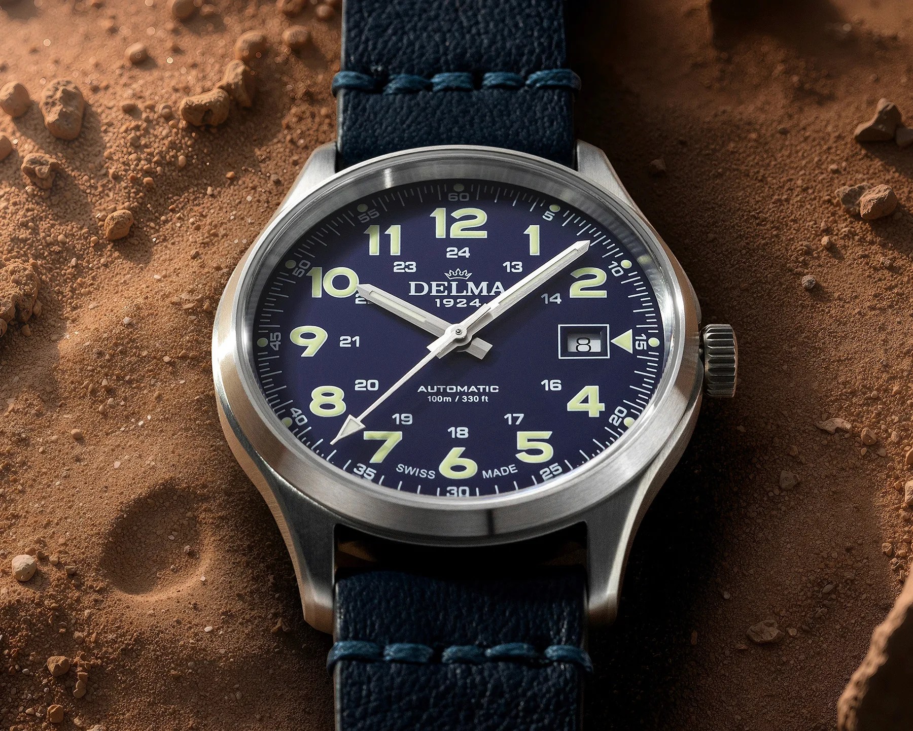 Close-up of a Delma 1924 automatic watch with a blue dial, luminous yellow numerals, and a blue leather strap on sandy ground.