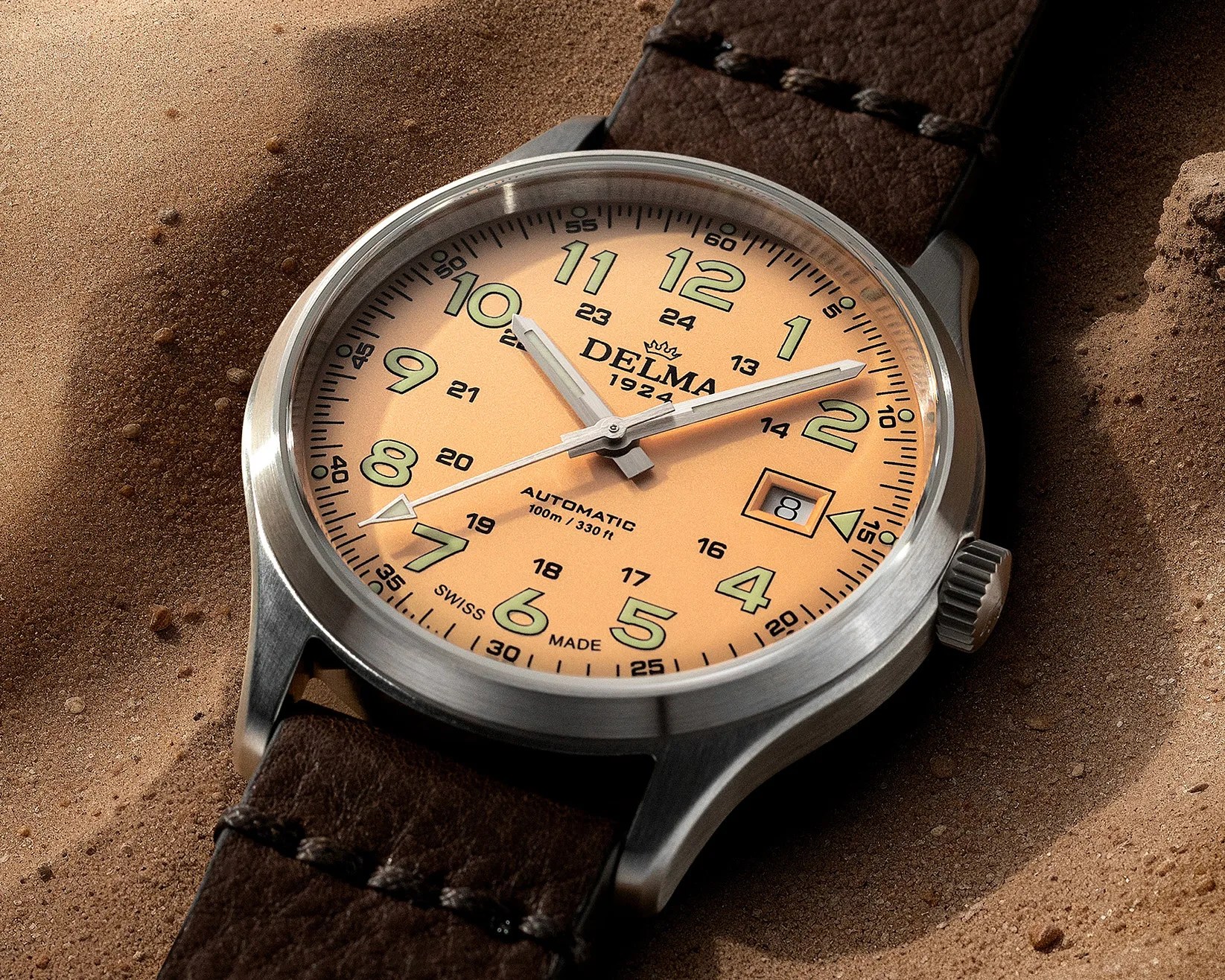 Close-up of a Delma automatic wristwatch with a beige dial, green numerals, date window, and brown leather strap on sand.