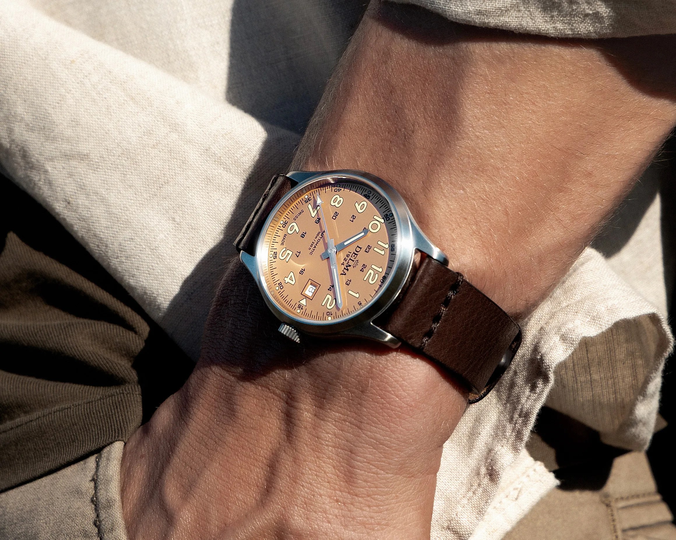 Wristwatch with a tan dial, silver case, and brown leather strap worn on a wrist over beige and brown fabric.