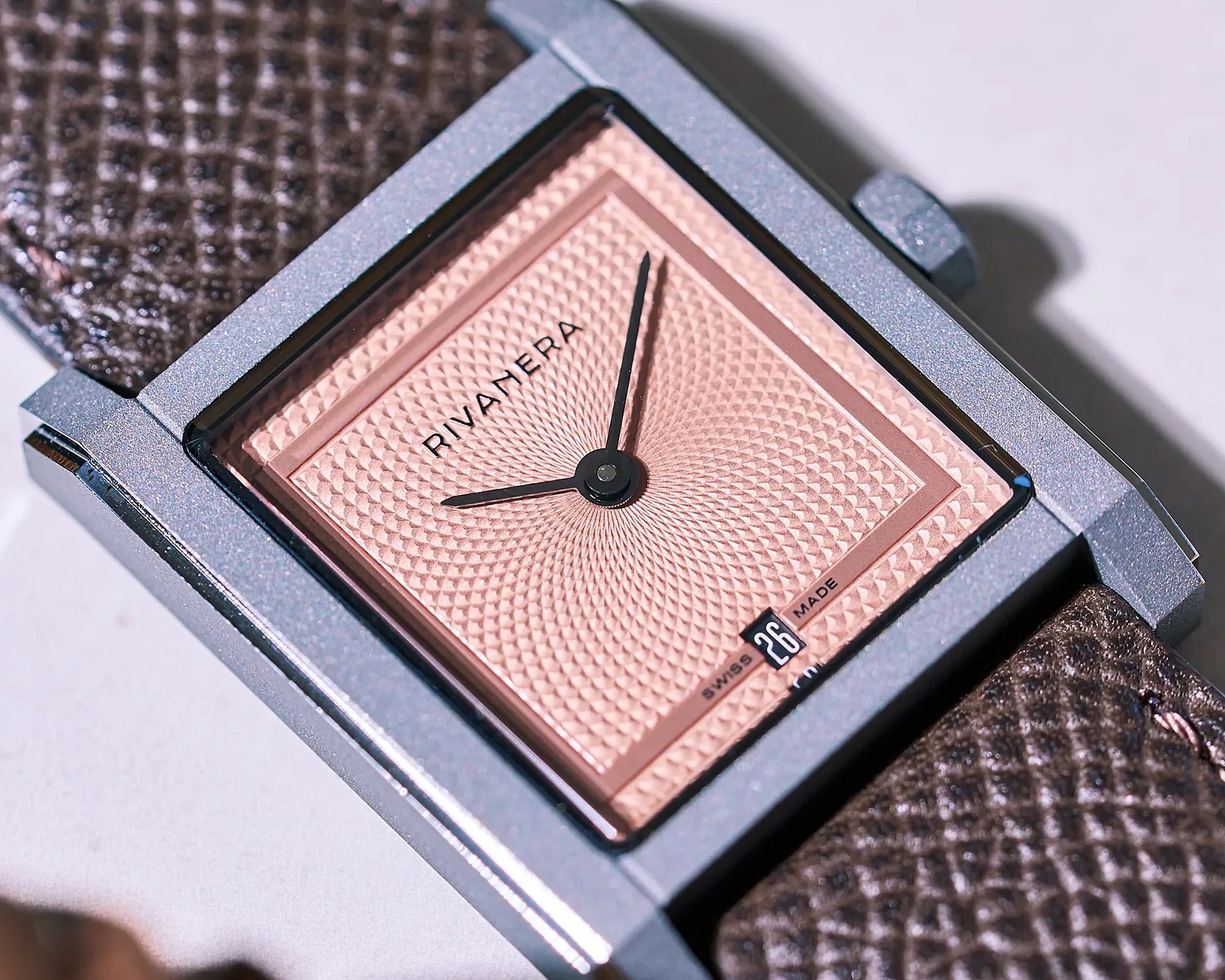 Square silver watch with textured pink dial and black hands on a brown textured leather strap.