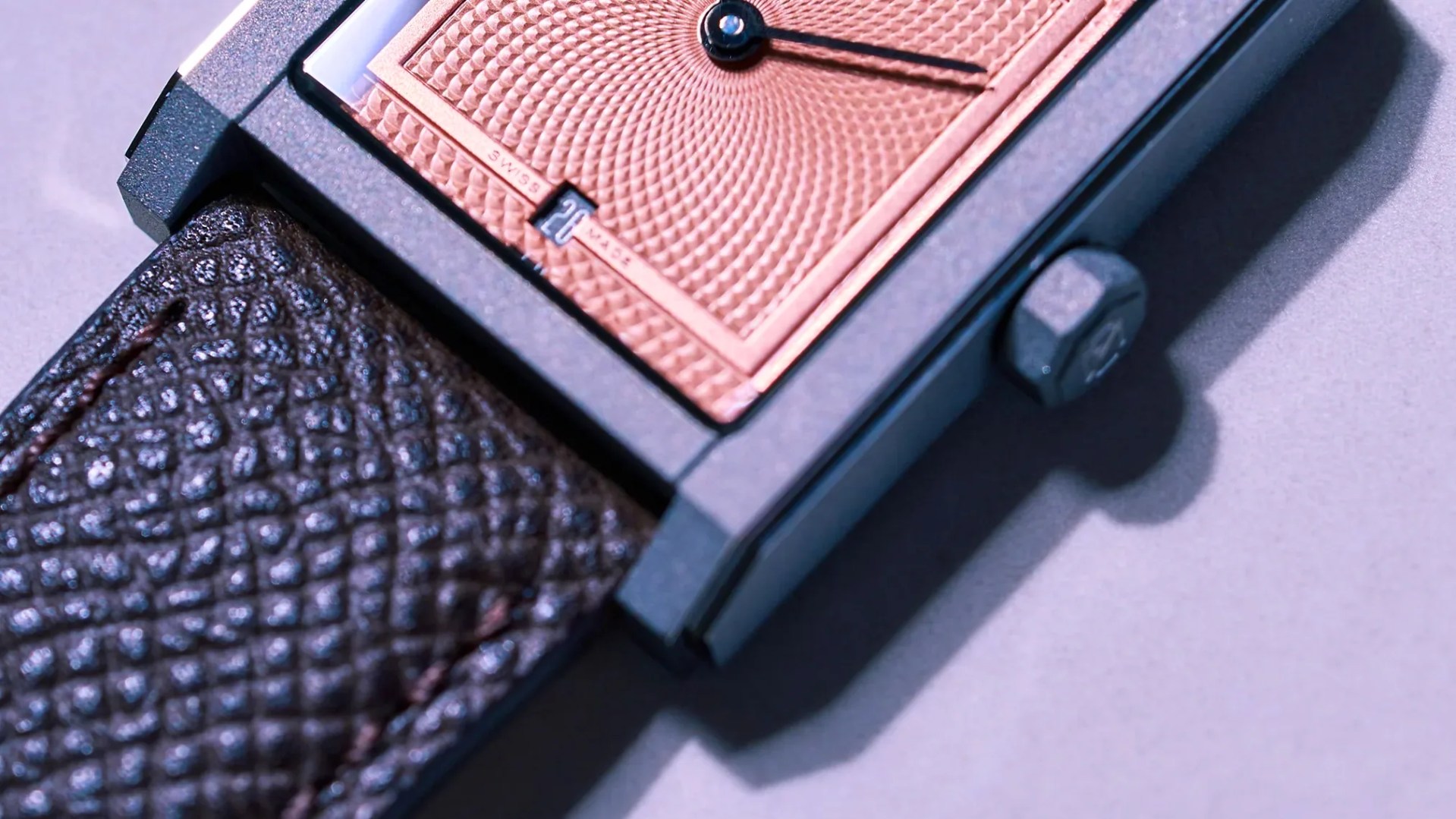 Close-up of a rectangular watch with a textured rose gold dial, black hands, and a brown textured leather strap.