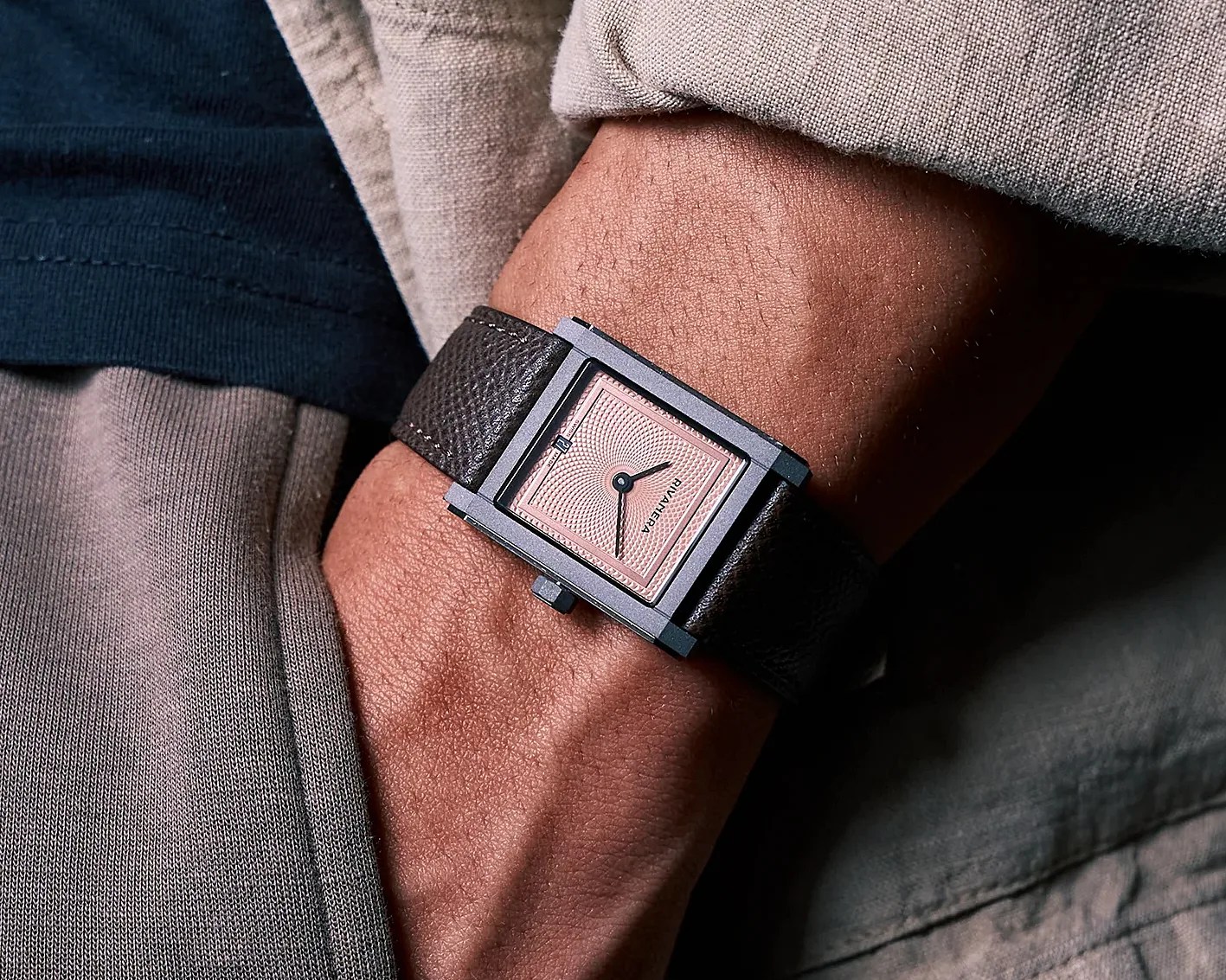 Rectangular wristwatch with a pink textured dial and black leather strap worn on a wrist.