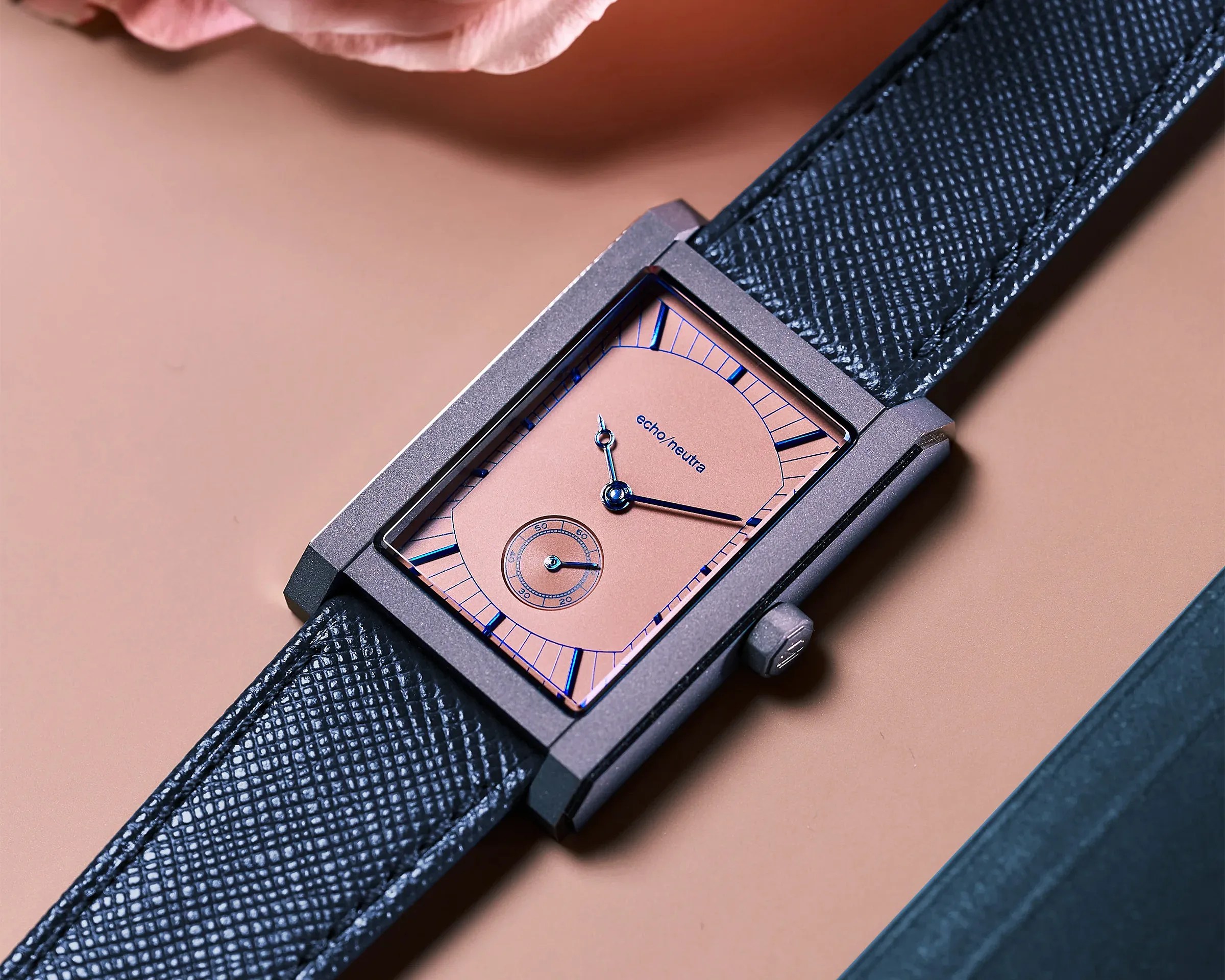 Rectangular wristwatch with a pink dial, blue hour markers and hands, and a textured black leather strap.