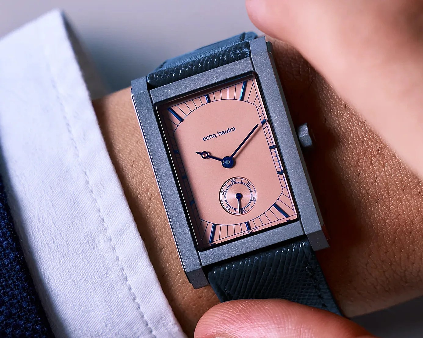 Rectangular wristwatch with a pink dial, blue hands, and black leather strap worn on a wrist.