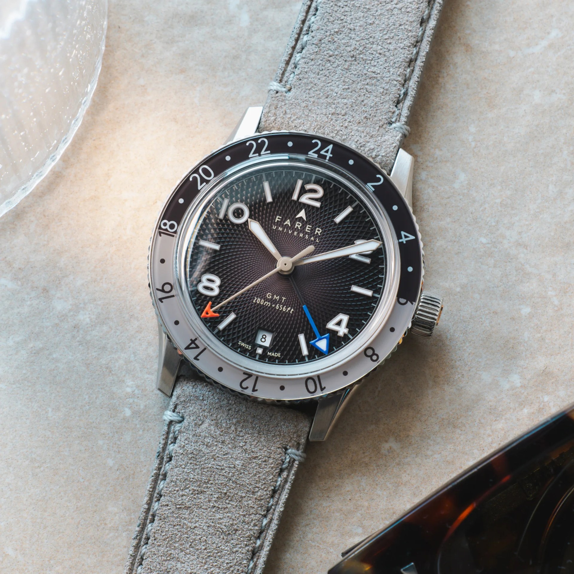 Close-up of a Farer Universal GMT watch with a textured black dial, silver case, and light gray suede strap on a beige surface.