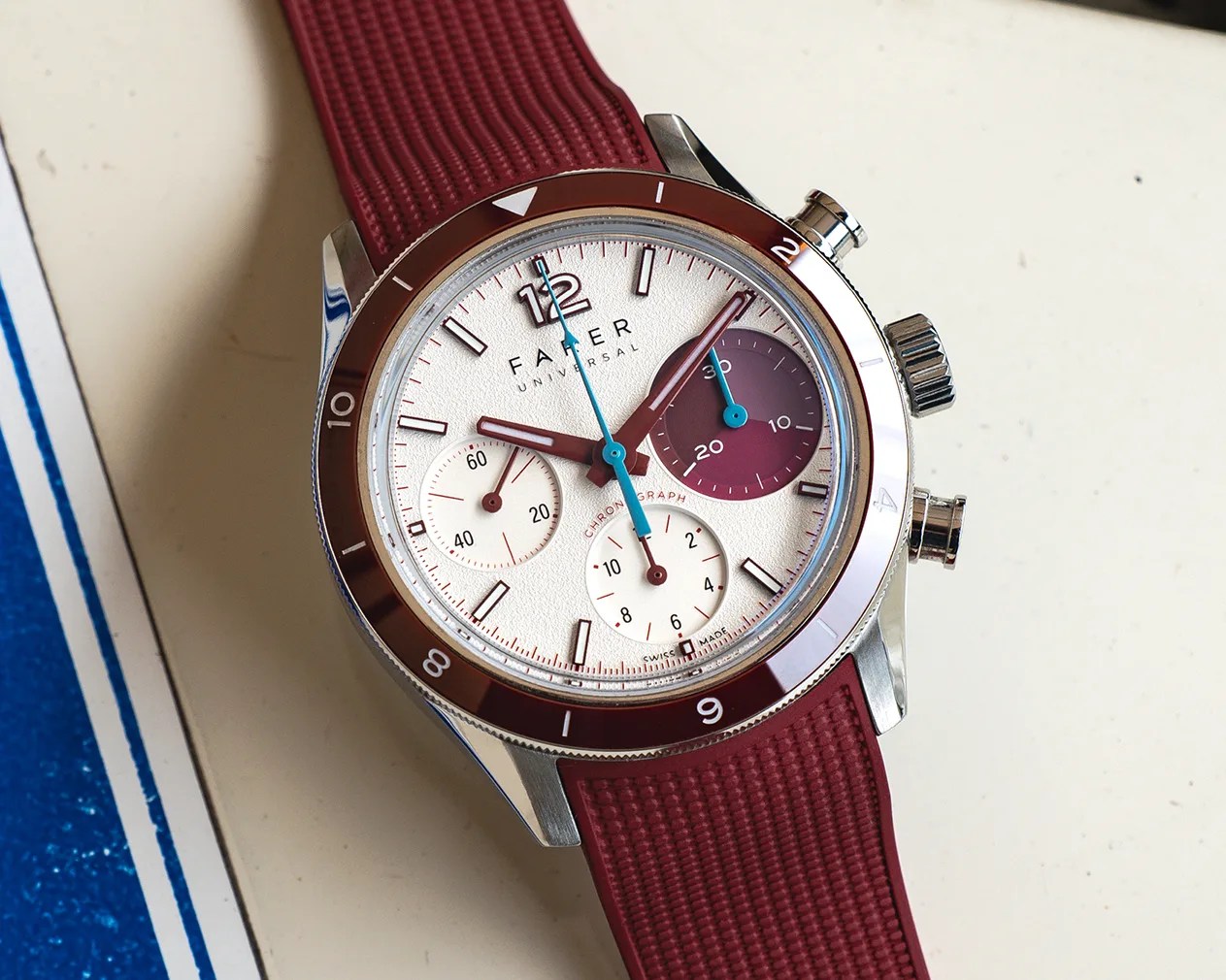 Silver chronograph watch with white and burgundy dial, blue second hand, and textured burgundy rubber strap.
