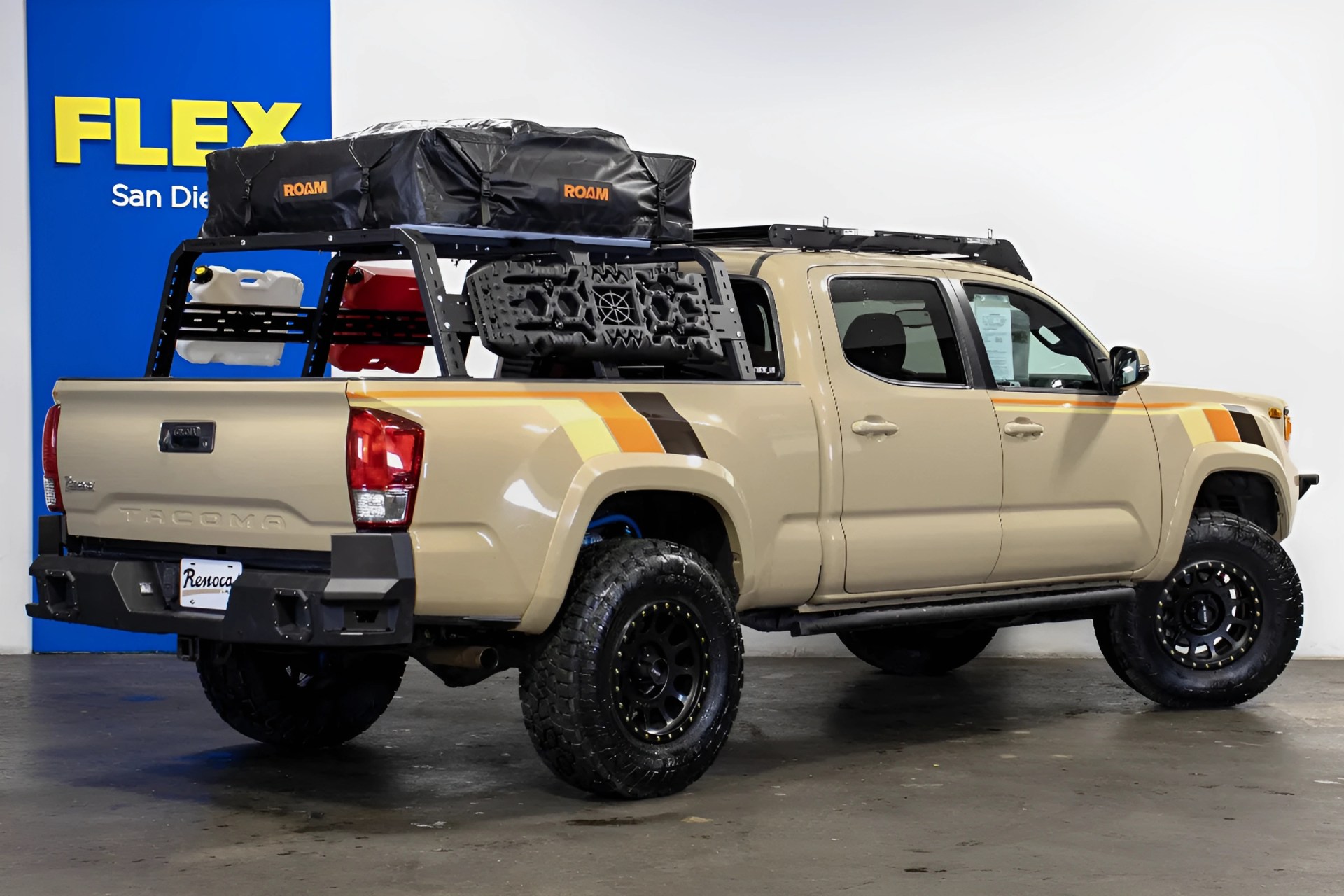 Beige Toyota Tacoma pickup truck with black off-road tires, roof rack, and camping gear in the bed.
