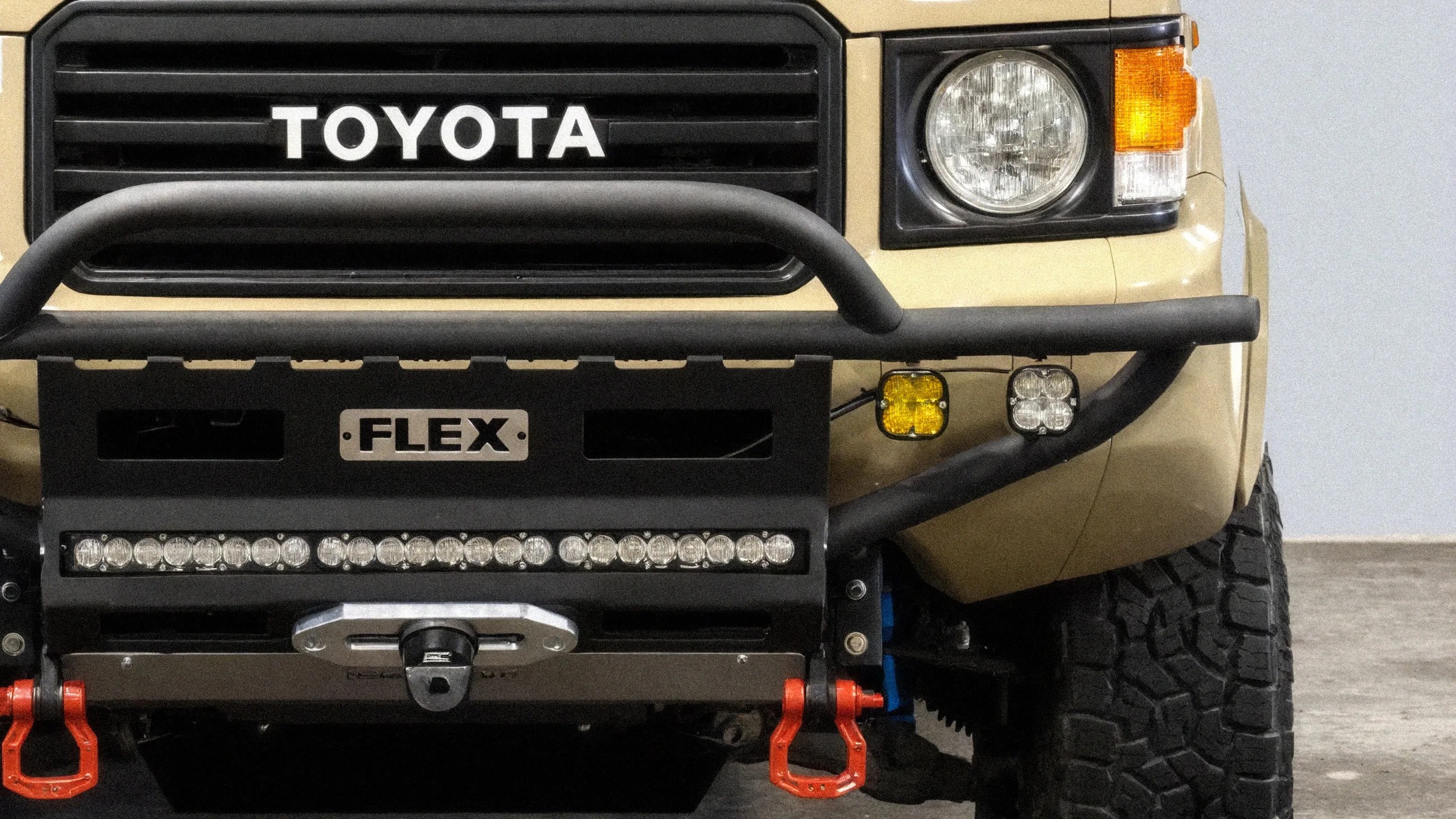 Front view of beige Toyota vehicle with black off-road bumper, LED light bar, and red tow hooks.