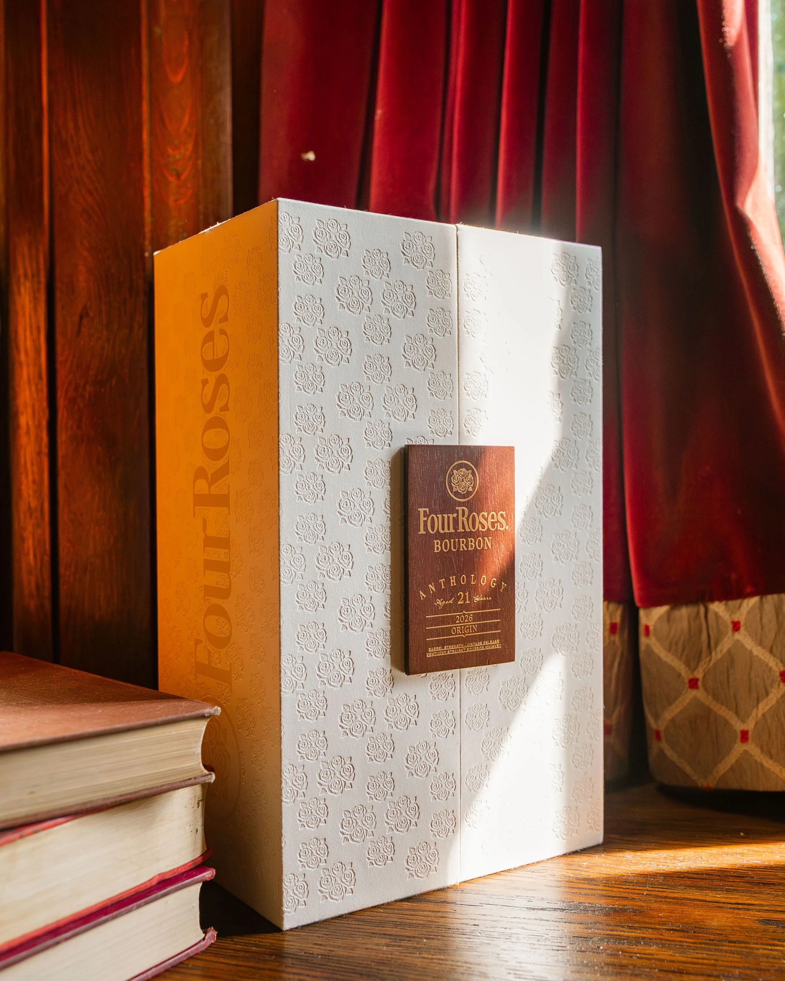 White and orange Four Roses Bourbon Anthology 21-year-old box with embossed rose patterns on a wooden surface near stacked books and red curtains.
