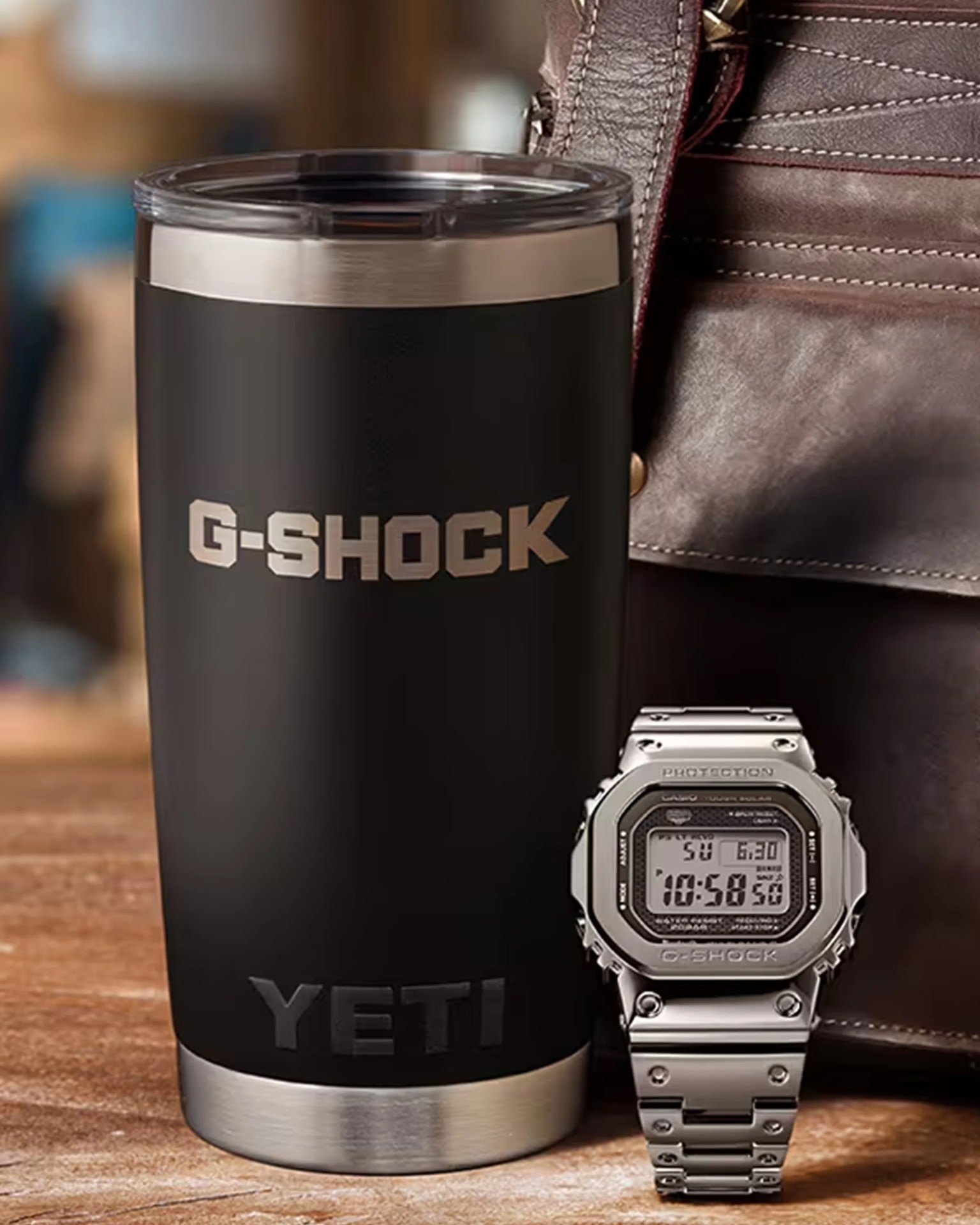 Black and silver YETI tumbler with G-SHOCK logo next to a silver metal G-SHOCK digital watch on a wooden surface.