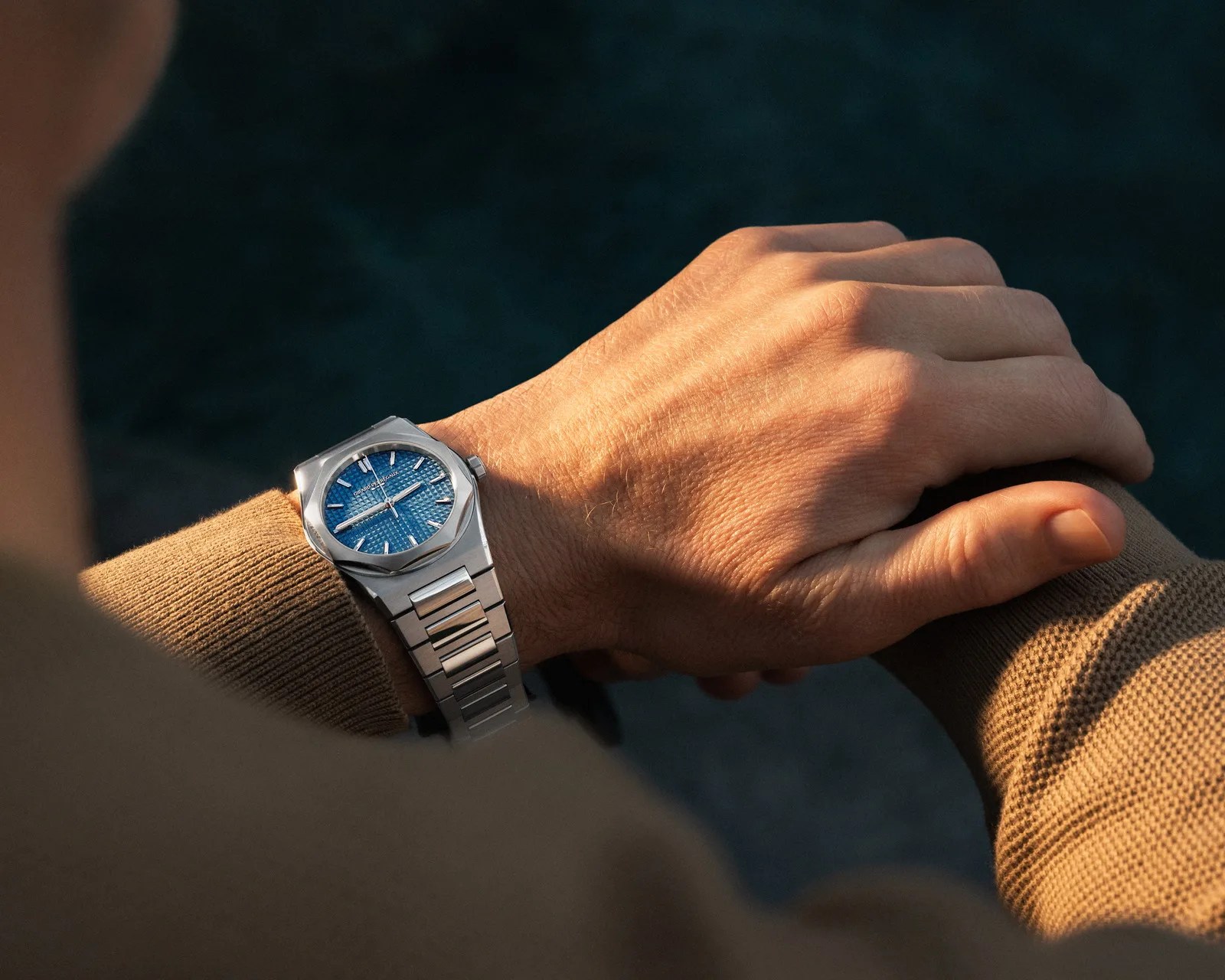 Silver metal wristwatch with a textured blue dial worn on a wrist with a brown sweater.