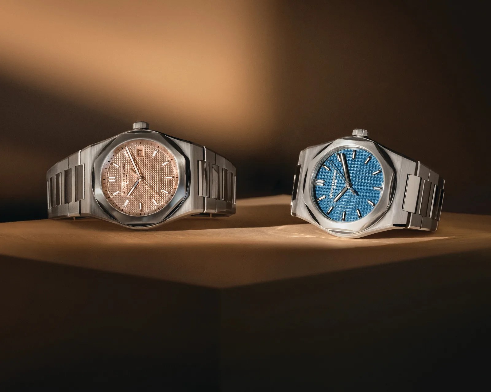 Two Girard-Perregaux stainless steel watches with textured dials, one pink and one blue, on a beige surface.