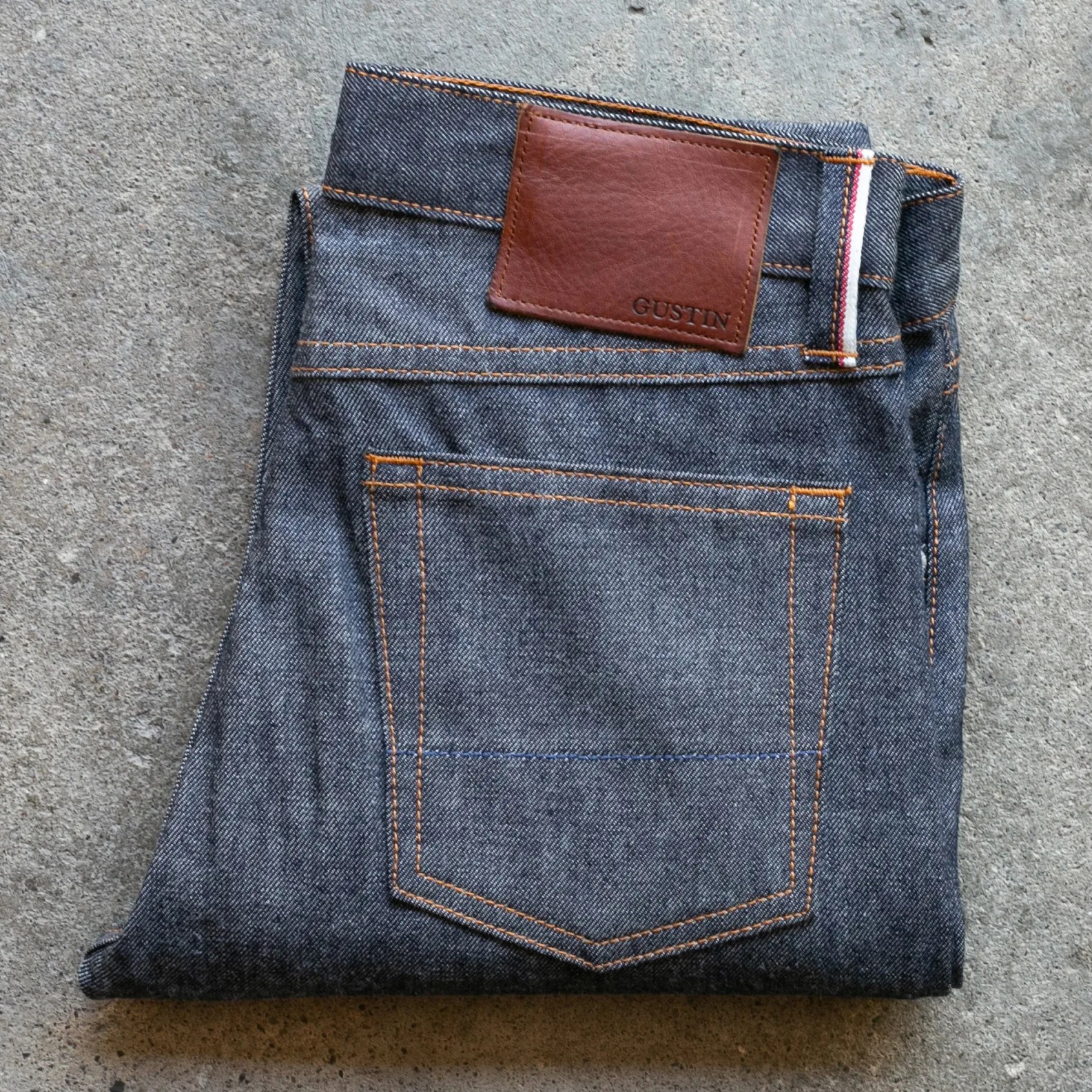 Folded dark blue denim jeans with orange stitching and a brown leather patch labeled "GUSTIN" on a concrete surface.