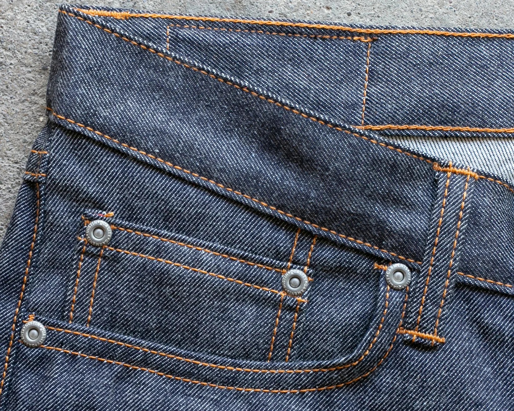 Close-up of dark blue denim jeans pocket with orange stitching and metal rivets on a concrete surface.