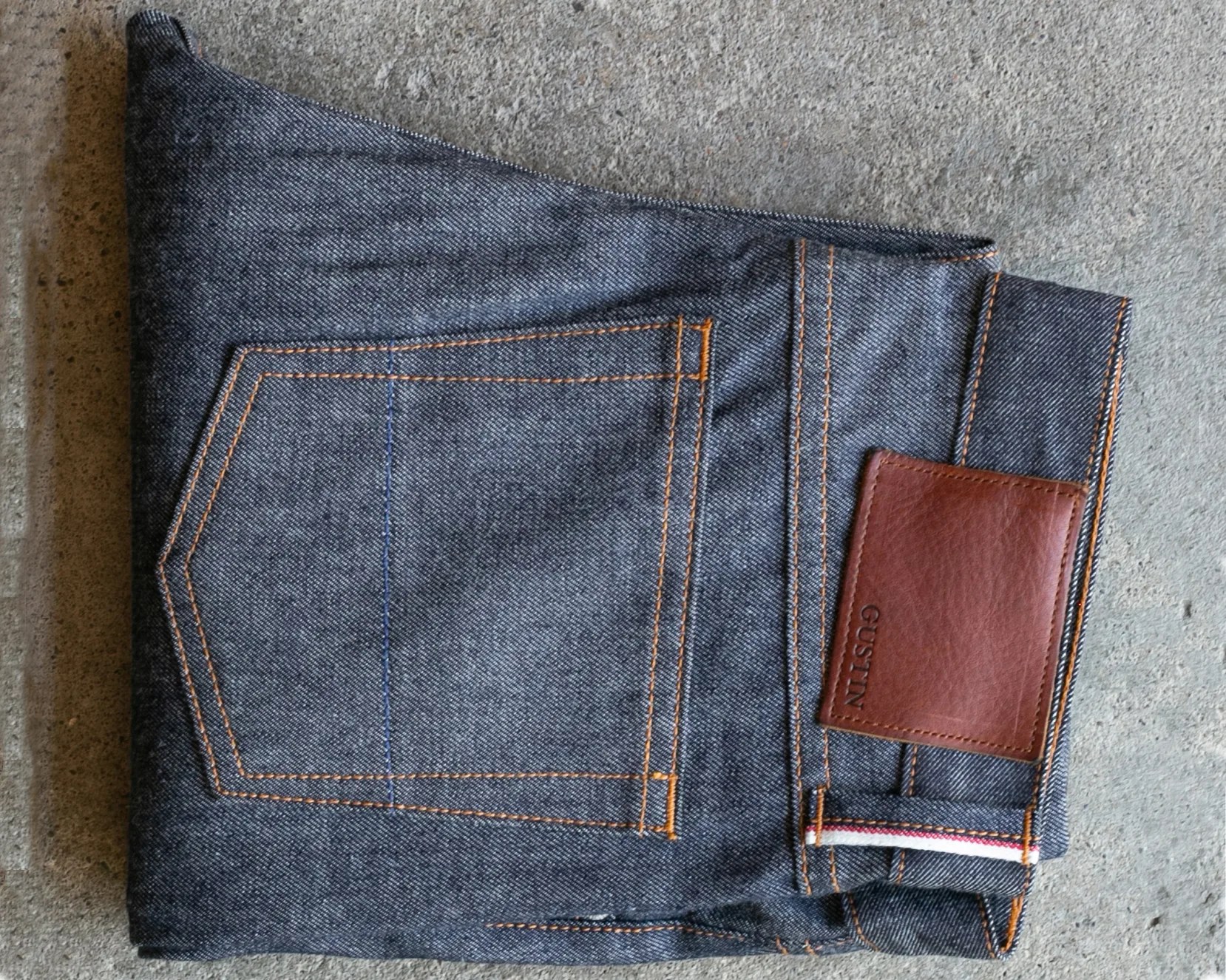 Folded dark blue denim jeans with orange stitching and a brown leather patch on a concrete surface.