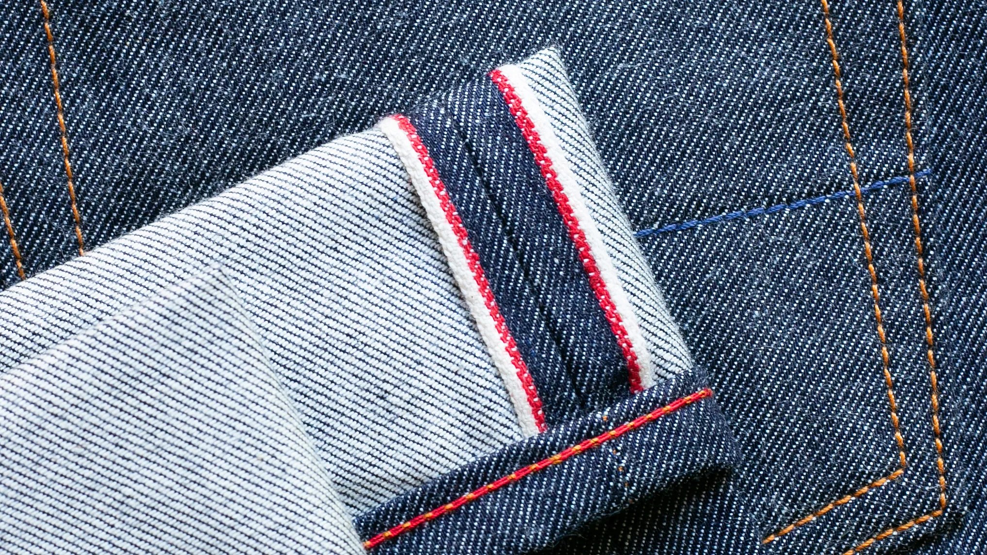 Close-up of dark blue denim fabric with orange stitching and a folded edge showing white and red selvedge detail.