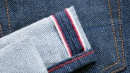 Close-up of dark blue denim fabric with orange stitching and a folded edge showing white and red selvedge detail.