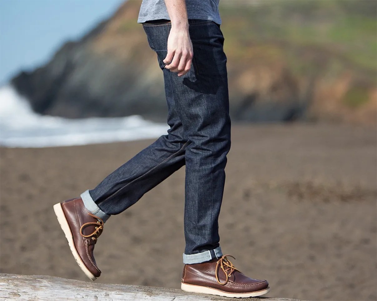 Person wearing dark blue rolled-up jeans and brown leather moccasin boots walking on a log at the beach.