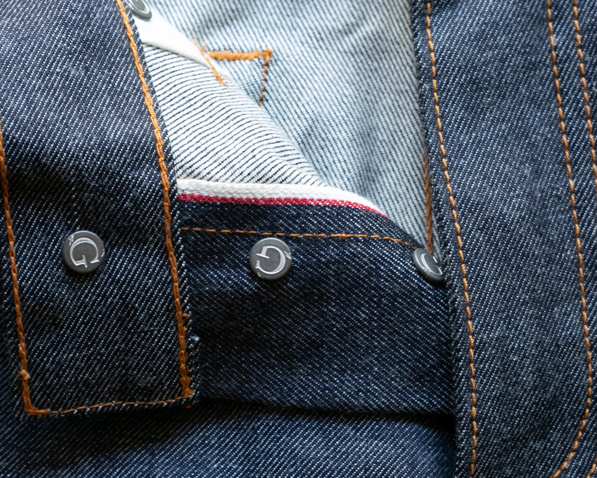 Close-up of dark blue denim fabric with orange stitching and metal buttons, showing the inside seam with a red and white selvedge edge.