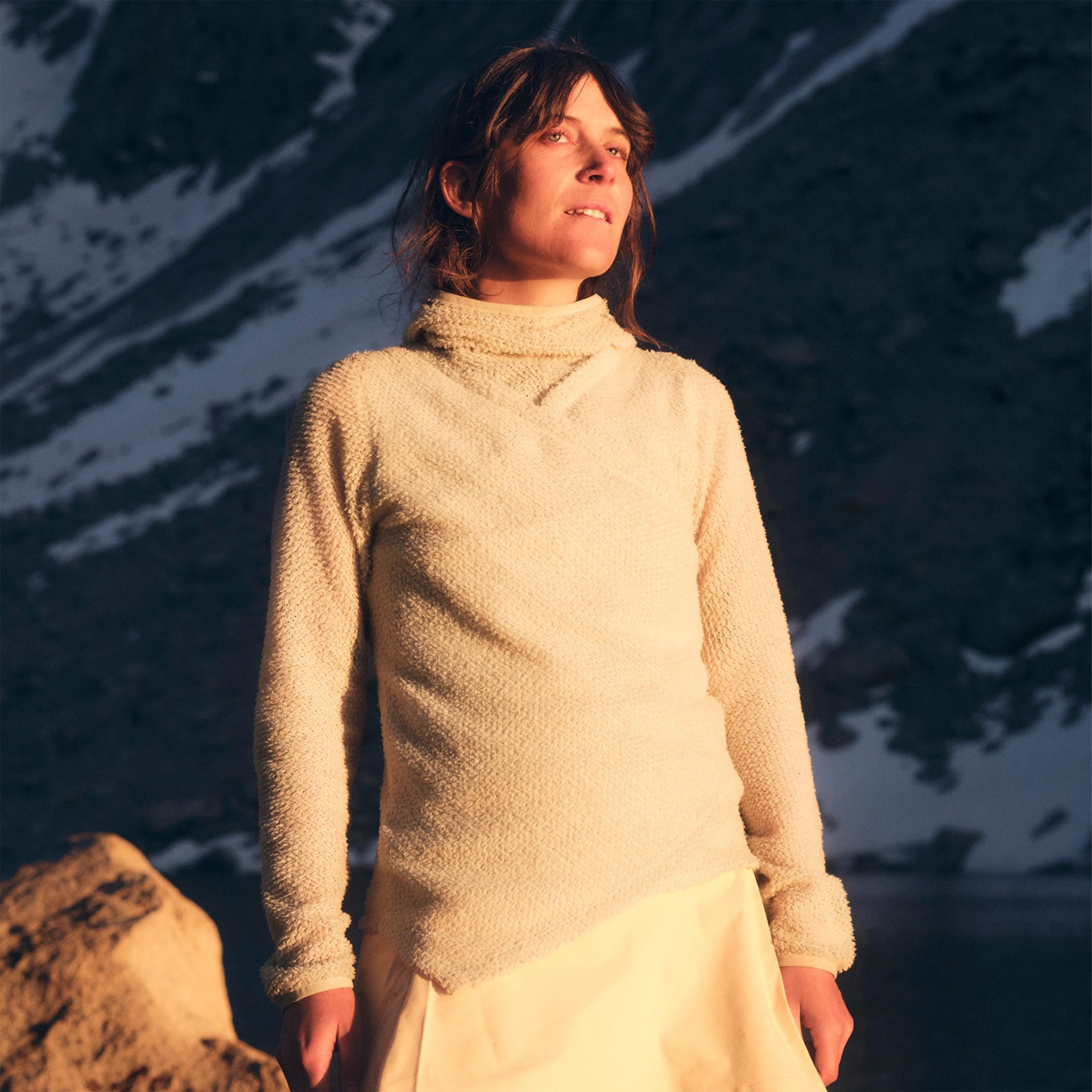 Cream-colored textured knit sweater with a high collar worn outdoors near rocky terrain.