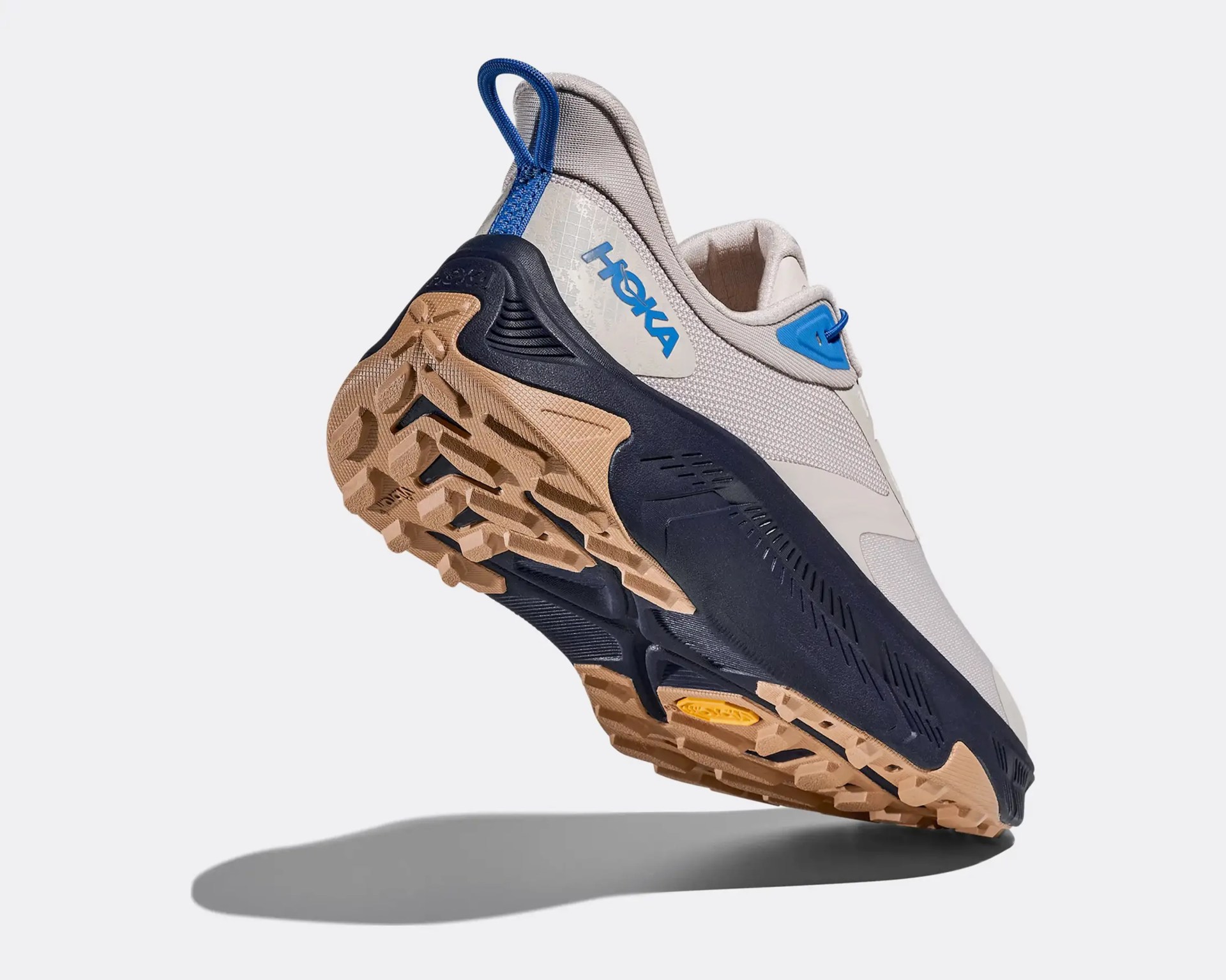 Beige and navy Hoka sneaker with blue accents and rugged tan outsole shown from the rear bottom angle.