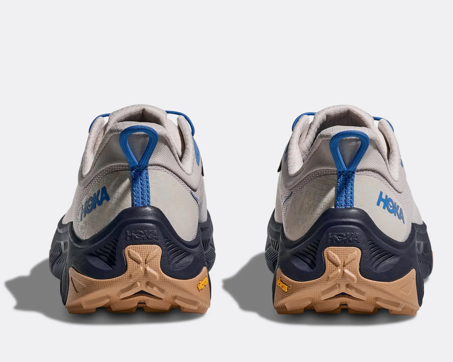 Pair of beige and navy Hoka trail running shoes viewed from the back showing blue pull tabs and tan Vibram soles.