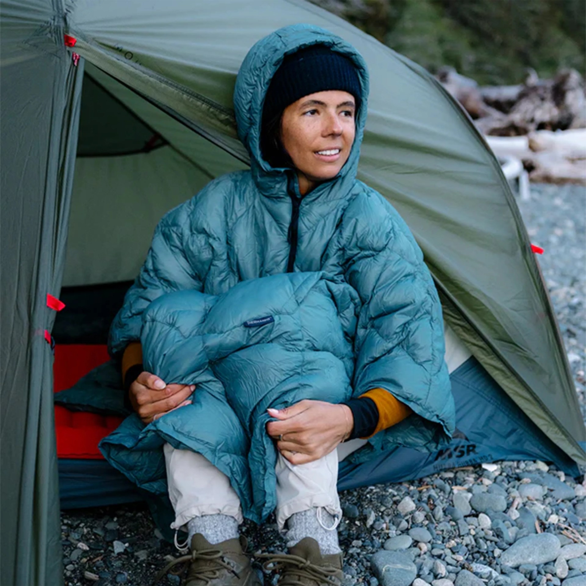 Person wearing a teal insulated hooded jacket and black beanie sitting at the entrance of a green tent on rocky ground.