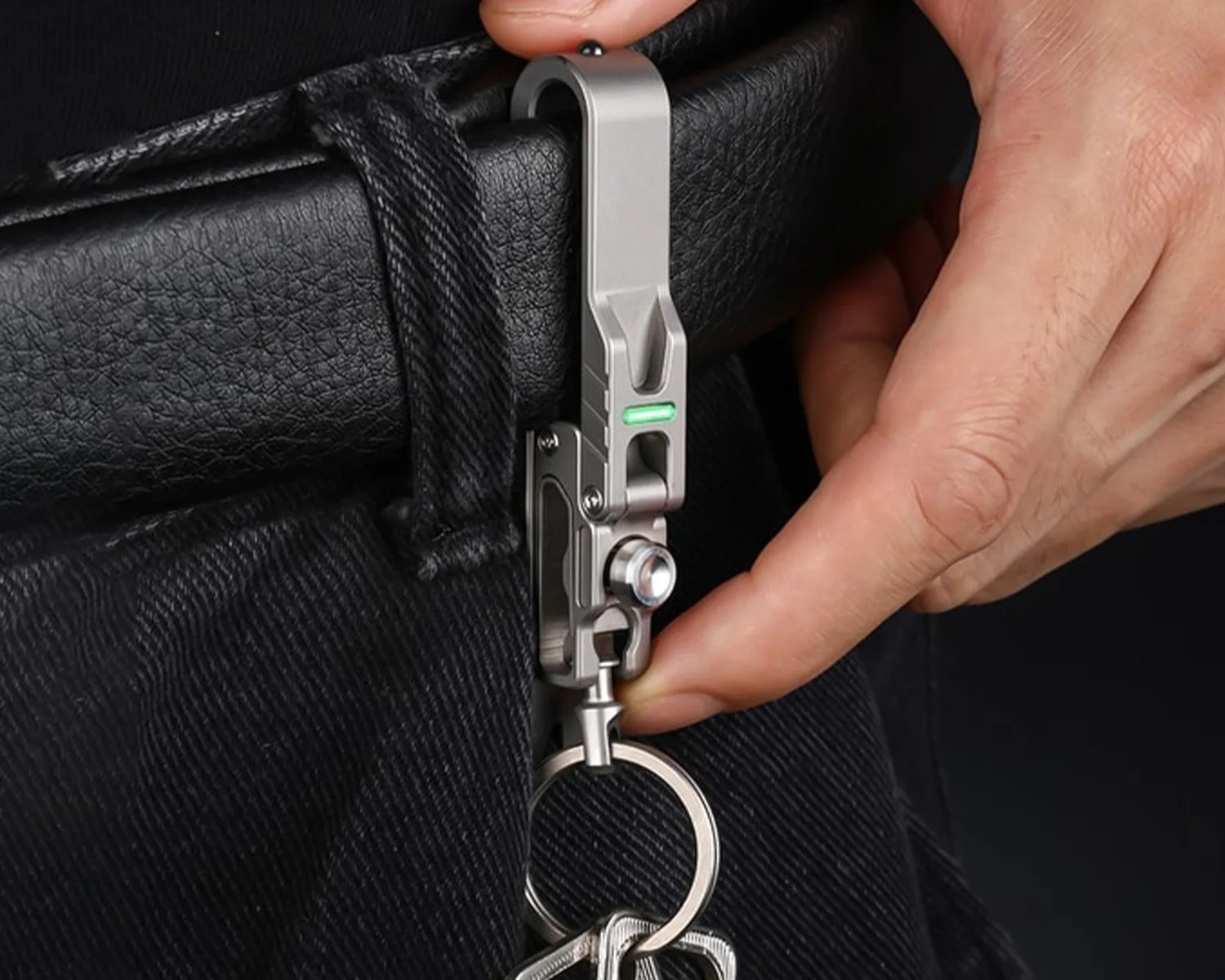 Metal keychain clip with a green indicator light attached to a black belt loop, held by a person's hand.