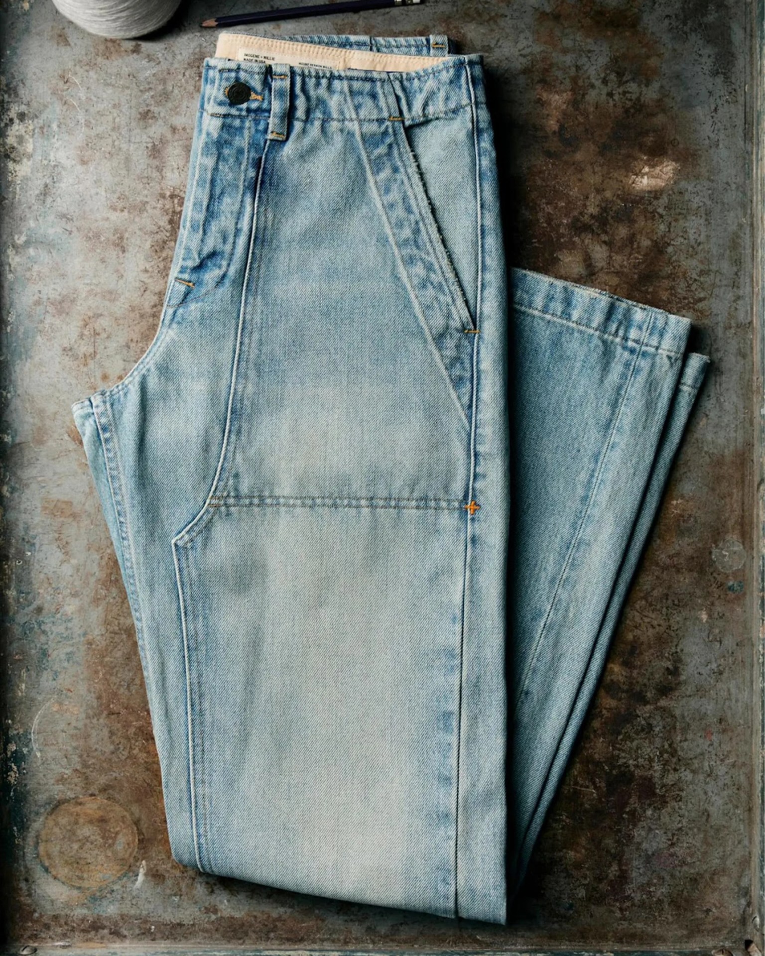 Light blue denim jeans with visible stitching and a button on a textured metal surface.