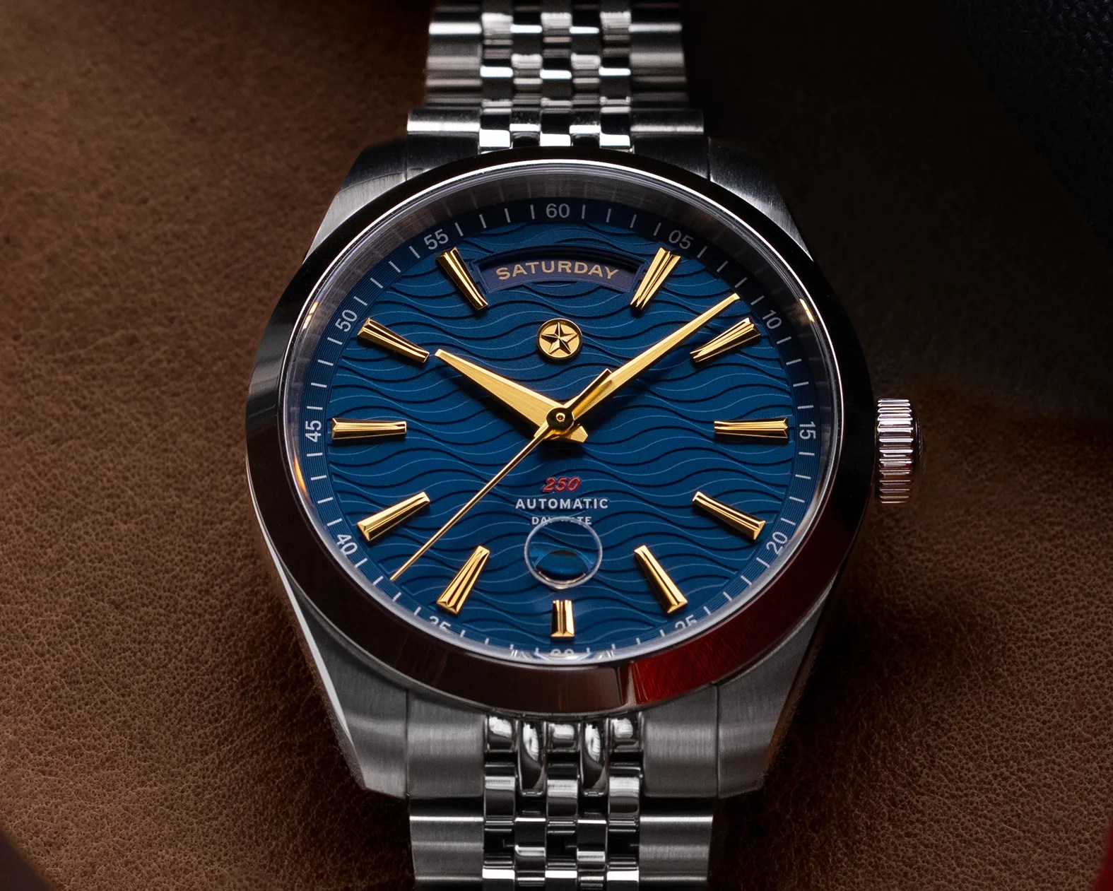 Stainless steel wristwatch with blue wave-patterned dial, gold hour markers and hands, and day-date display.